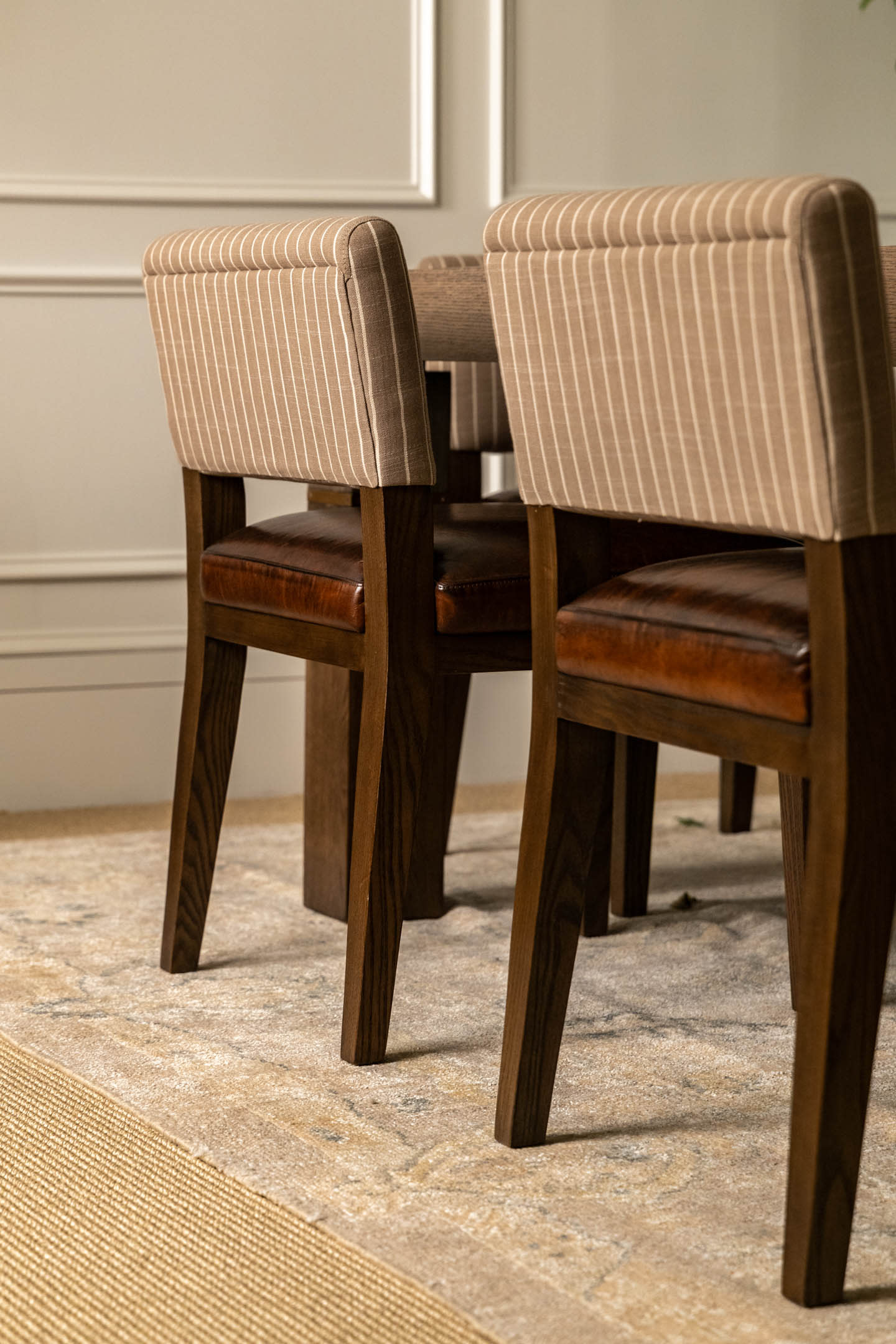 Wooden dining chairs with striped beige upholstery and leather seats are placed on a textured rug against a white paneled wall, conveying elegance.