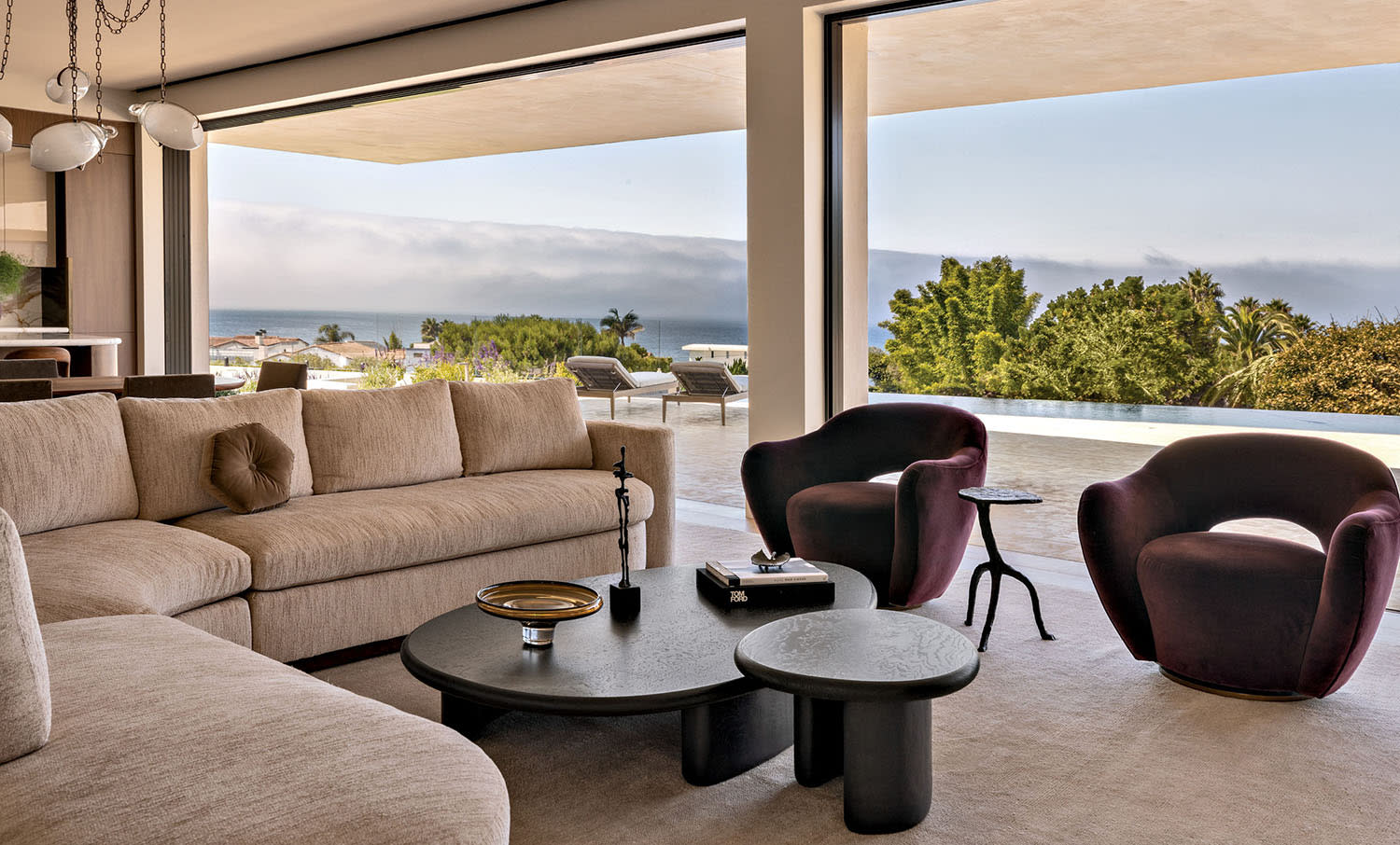 A tan curved sofa faces two dark armchairs alongside large windows looking towards an outdoor patio.