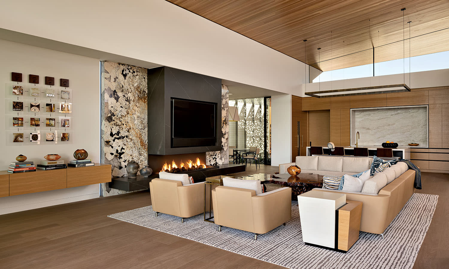 Luxurious modern living room with beige sofas, a central coffee table, and a wall-mounted TV above a sleek fireplace. A large window illuminates the space.