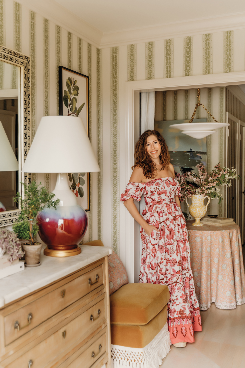 Portrait of designer Elizabeth Stamos leaning against a wall in a stripped paper and next to a small table covered with a pink patterned cloth.