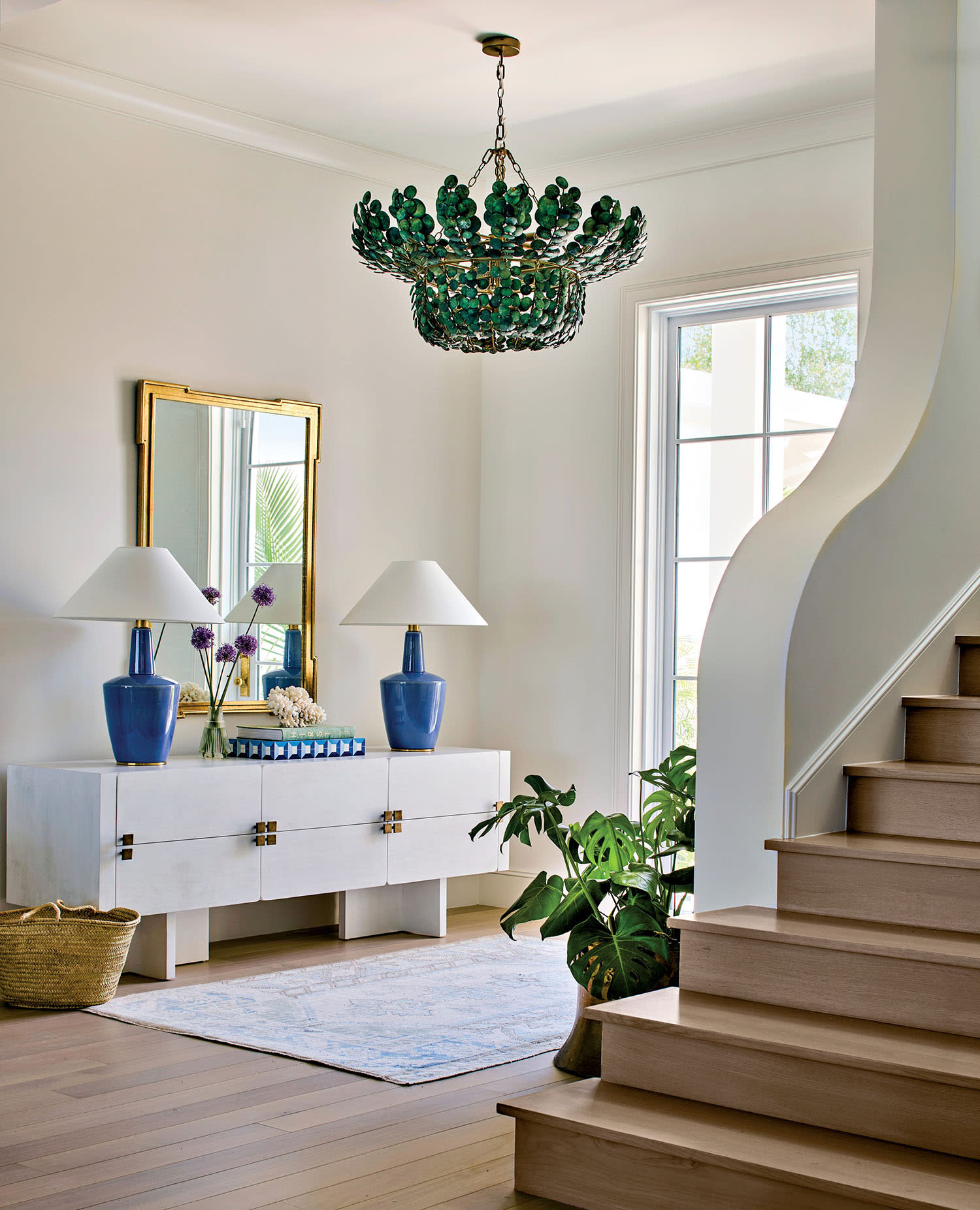 Bright entryway with a green glass chandelier, modern white sideboard, and blue lamps. A staircase curves elegantly beside leafy plants. Airy and inviting.