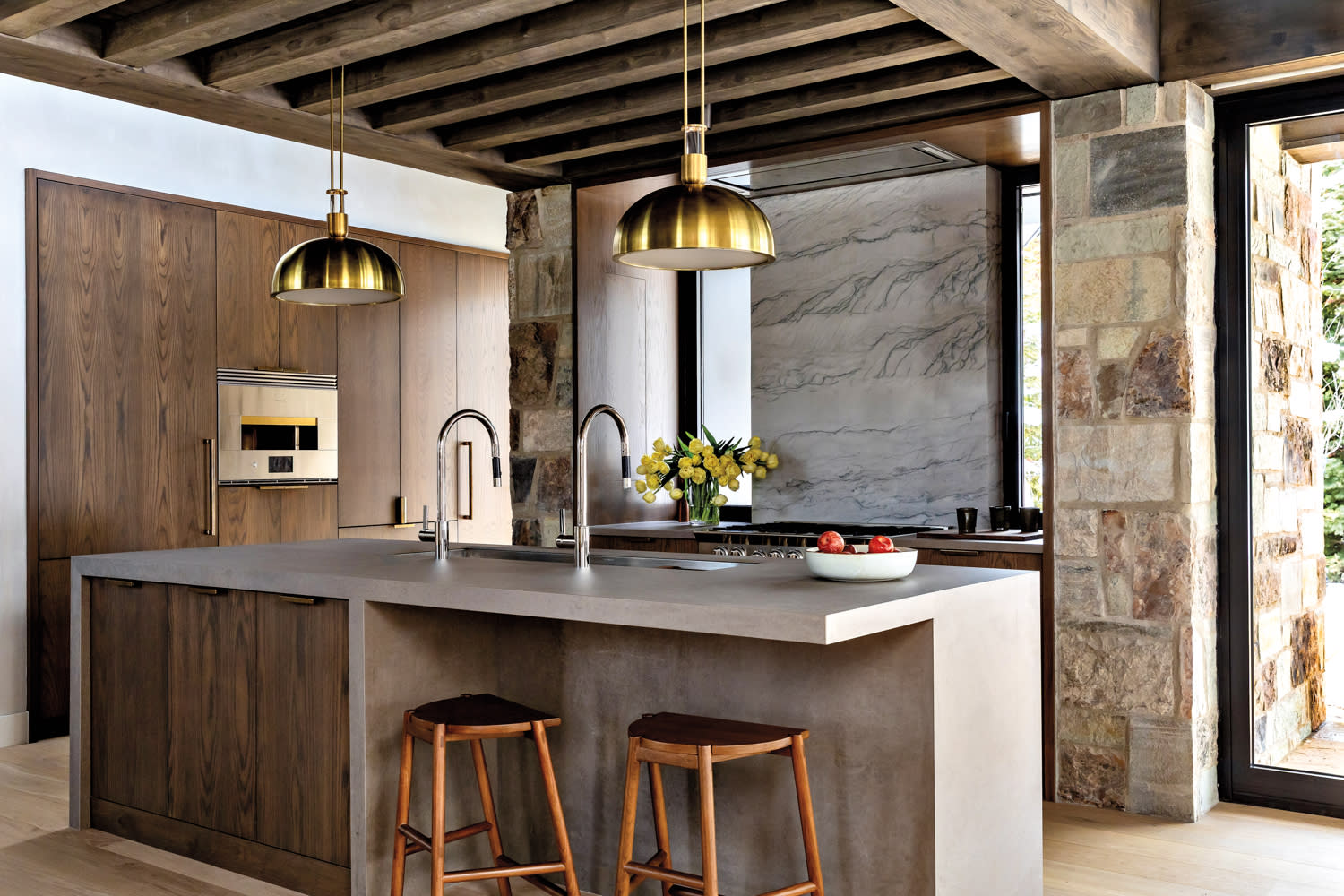 Modern kitchen with large island, two wooden stools, and brass pendant lights. Stone walls with wide windows create a warm, rustic ambiance.