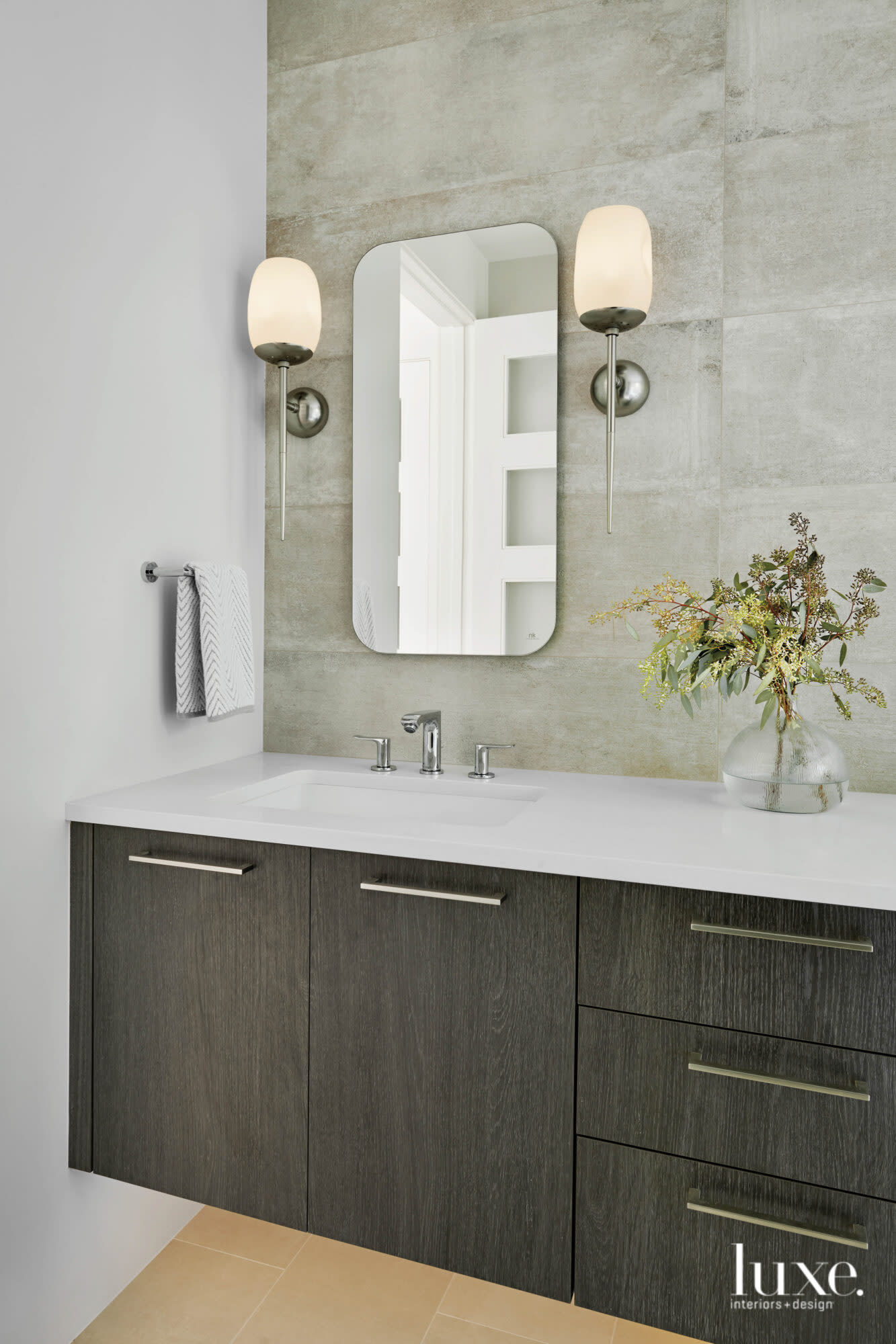 The primary bathroom features stone-like tile, a curvy mirror and a pair of shapely scones.