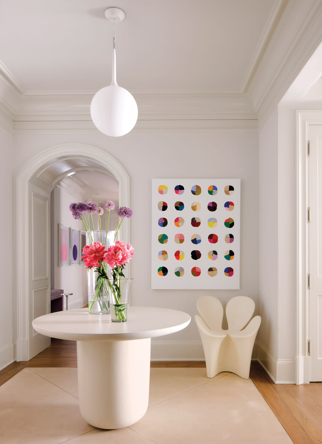 A minimalist room features a round table with vibrant pink and purple flowers in a vase. A colorful dot painting is on the wall, under a modern white pendant light.