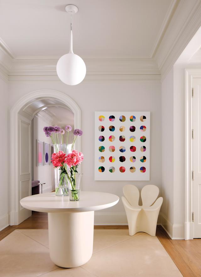 A minimalist room features a round table with vibrant pink and purple flowers in a vase. A colorful dot painting is on the wall, under a modern white pendant light.