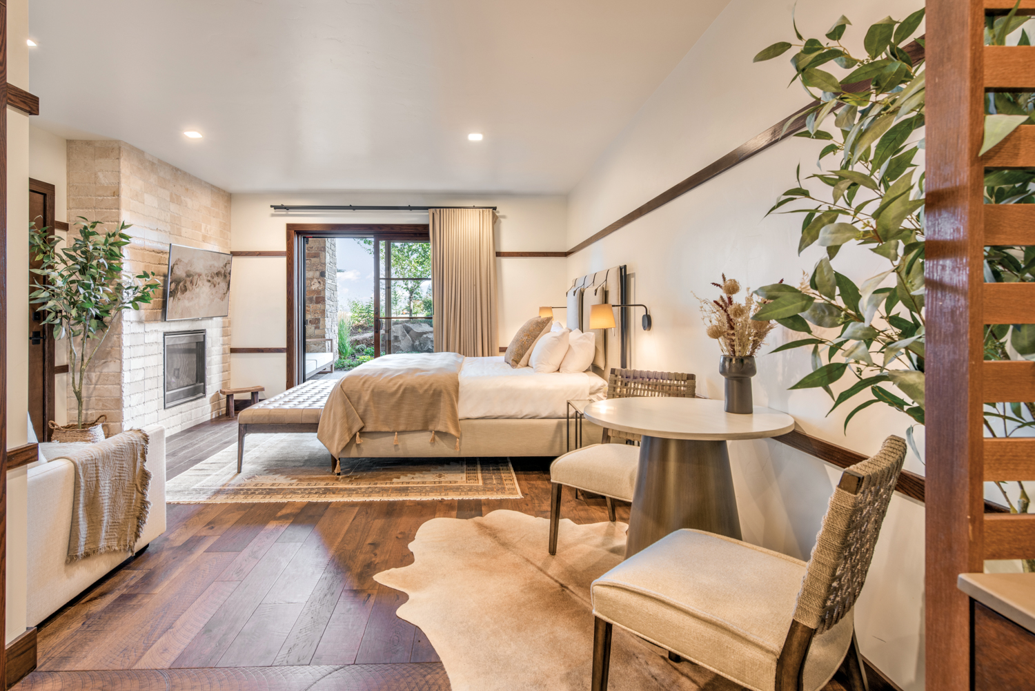 Bed with cream and white bedding faces stone fireplace and parallel to sliding door leading to a patio