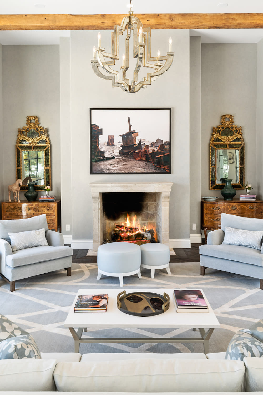 A stylish living room features two armchairs, a coffee table, a lit fireplace, and ornate wall mirrors. A large framed photo of a rustic boatyard hangs above the mantel. A chandelier hangs from the ceiling.