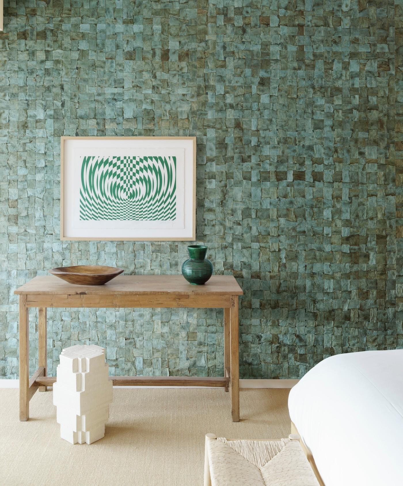 A rustic wooden table against a textured green wall holds a wooden bowl and green vase. Above hangs geometric art, with a cozy, minimalist vibe.