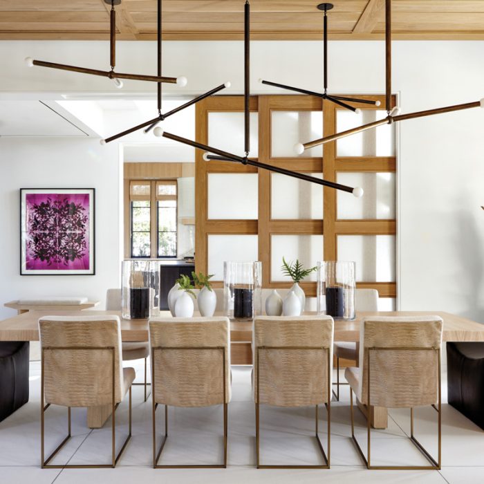 A colorful Wendy Small artwork in the foyer is visible from the dining area, which opens to the kitchen via paneled sliding doors. Apparatus' Arrow pendants seem to float over All Custom Cabinetry's white oak table, which is lined with armless Milo Baughman side chairs and capped by custom Moore & Giles leather ottomans. modern beach house