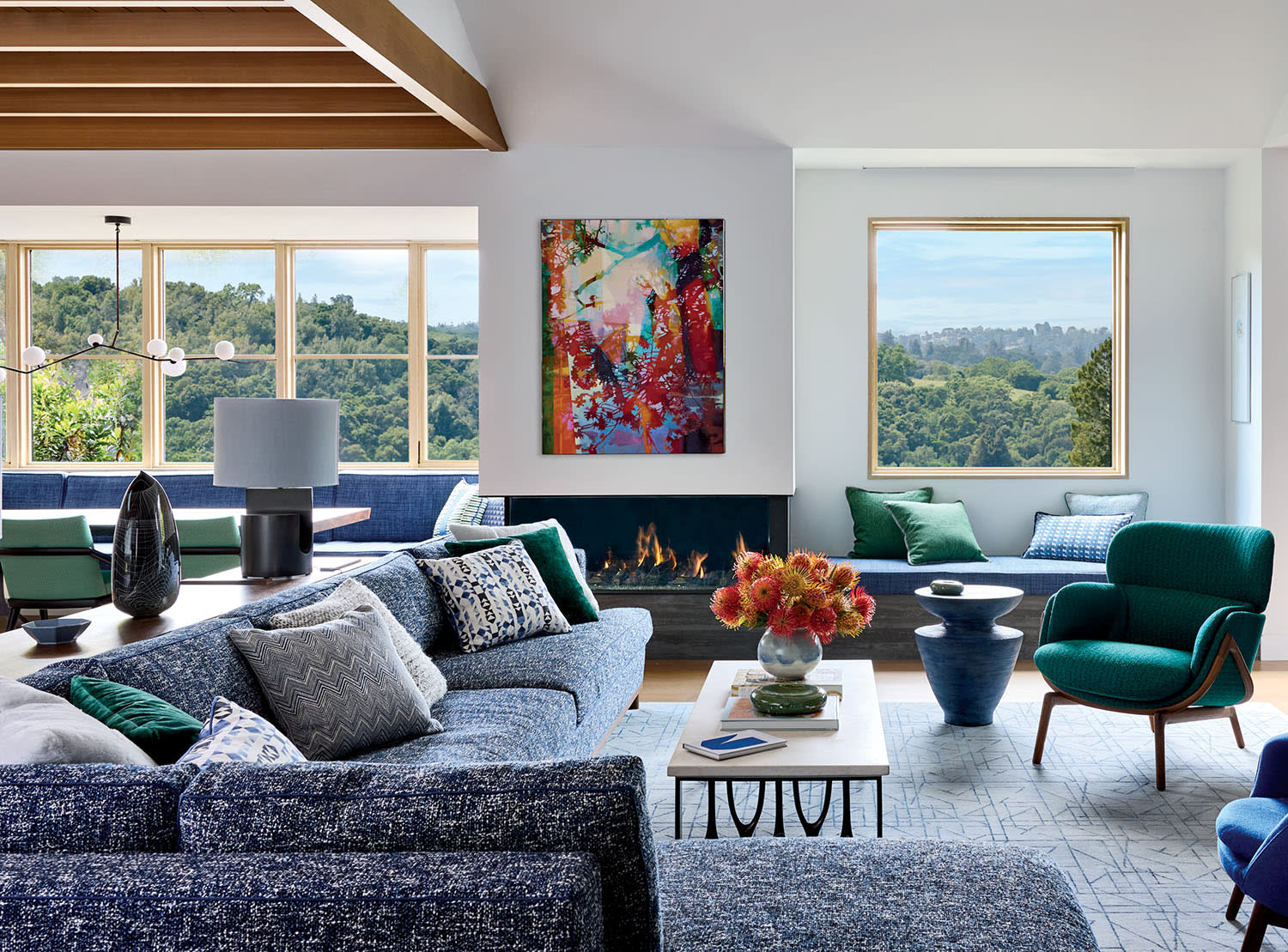 Stylish living room with large windows, blue and green armchairs, abstract art, and a modern chandelier. Light and airy with a cozy, inviting feel.
