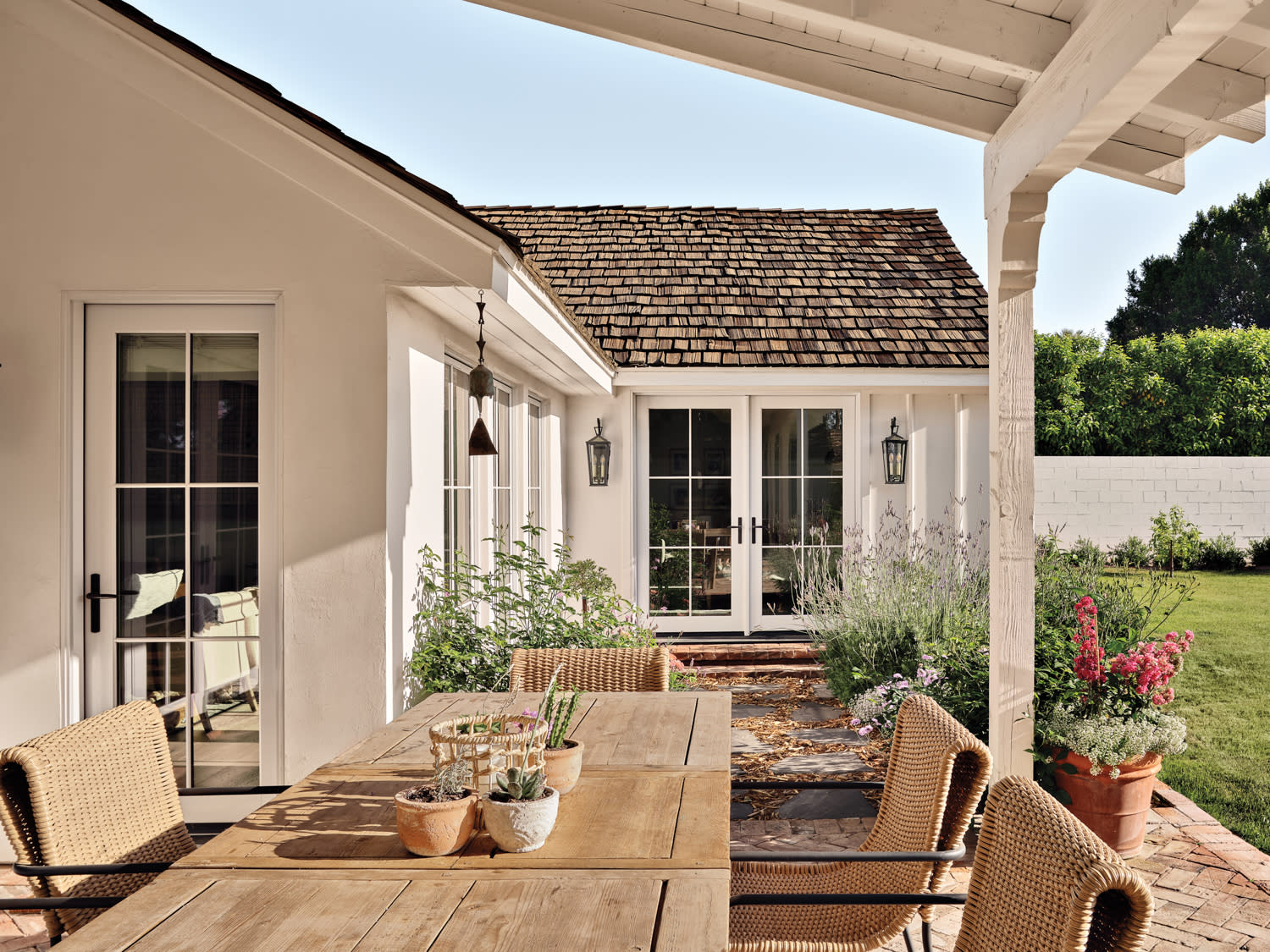 Wood dining table and woven chairs on the patio of a shingle-roofed farmhouse-style home