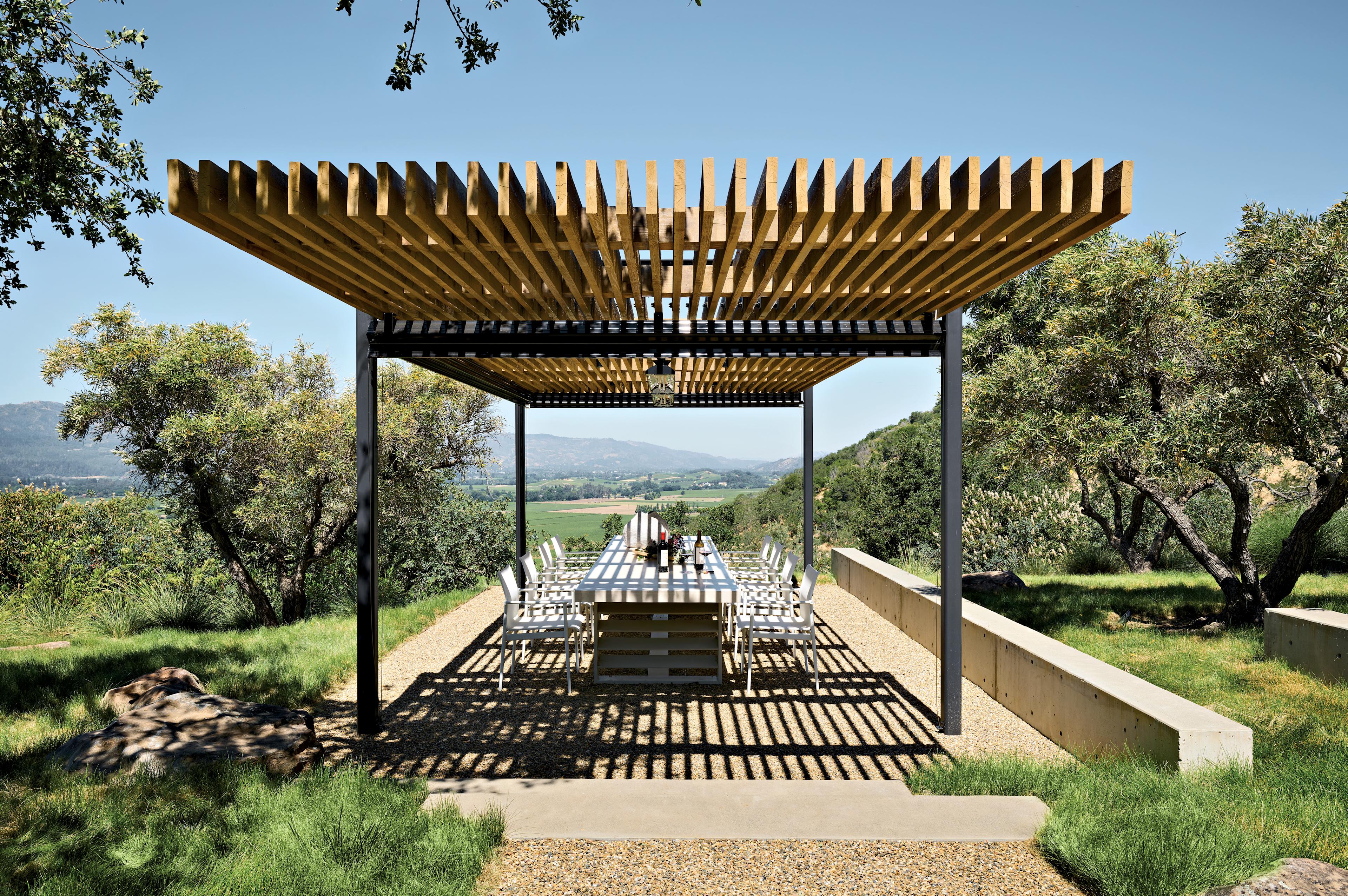A steel and cedar pergola sits over a table and chairs in an outdoor dining area flanked by trees.
