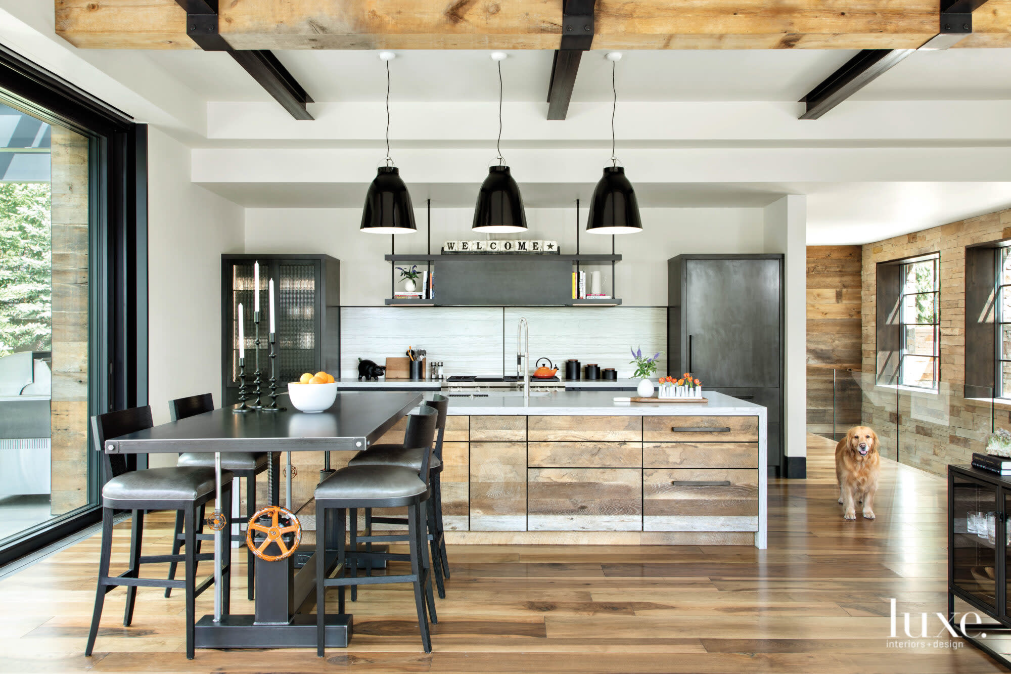 A kitchen features an adjustable metal table, an island with recycled wood and large black pendant lights.