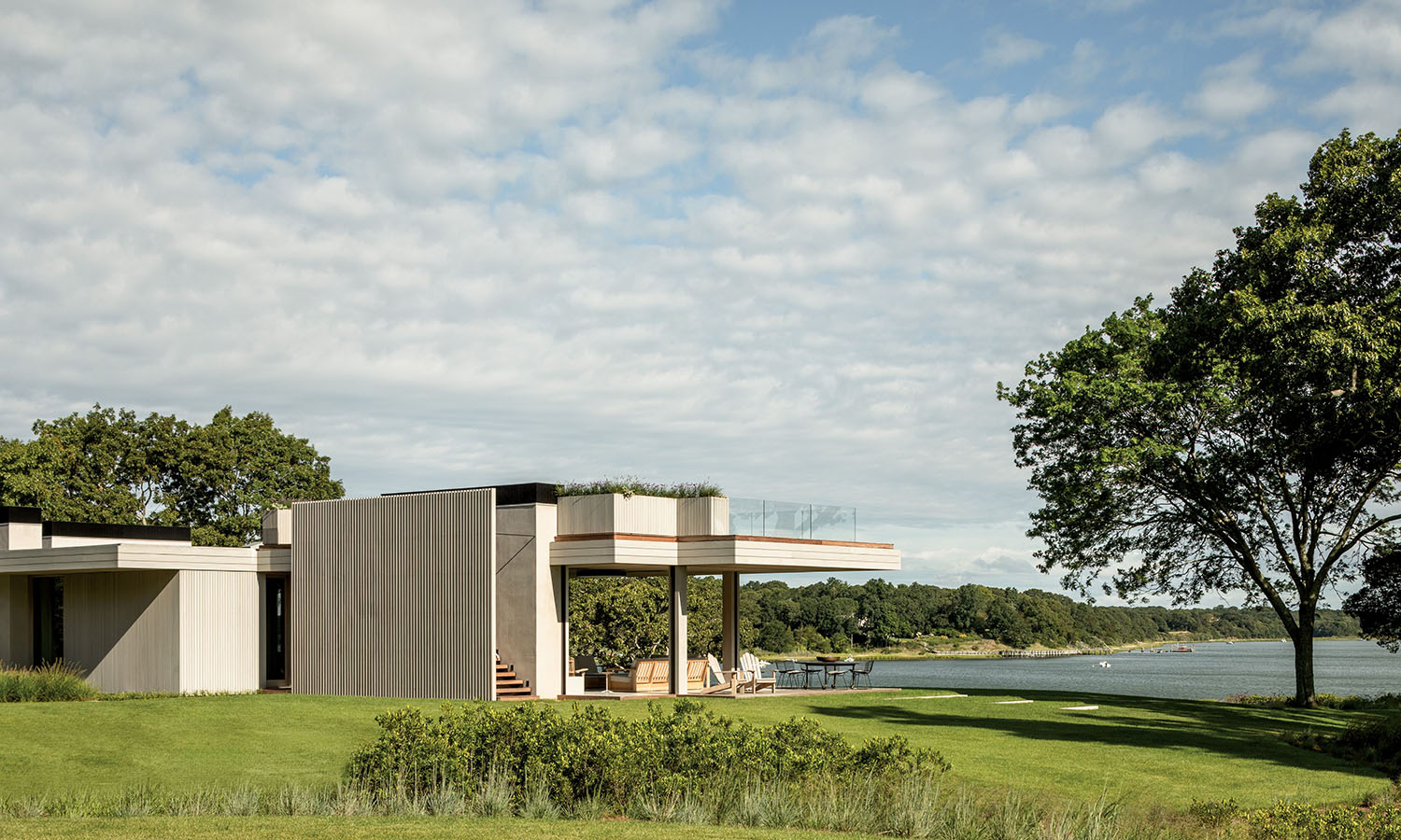 Modern house with flat roof set amidst a lush green lawn by a serene lake. The sky is partly cloudy, and a large tree adds natural charm to the scene.
