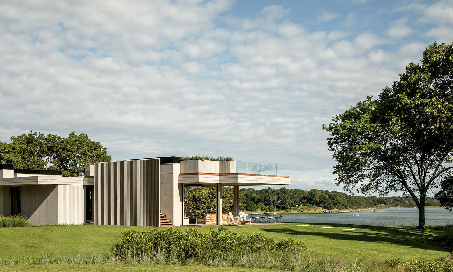 Modern house with flat roof set amidst a lush green lawn by a serene lake. The sky is partly cloudy, and a large tree adds natural charm to the scene.