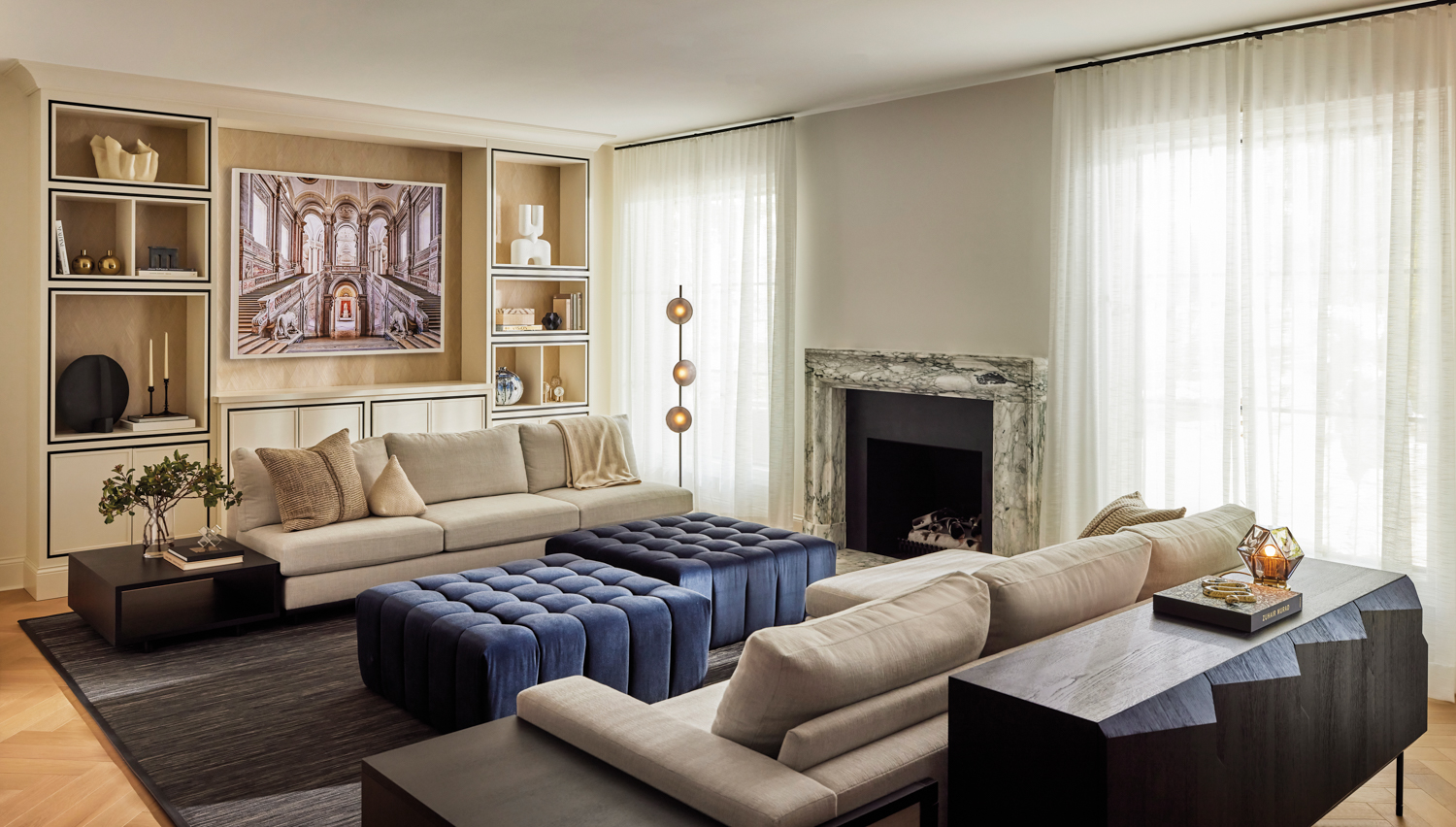 a family room with a photograph, a marble fireplace, low-back gray couches and two tufted, navy mohair ottomans by Marli Jones