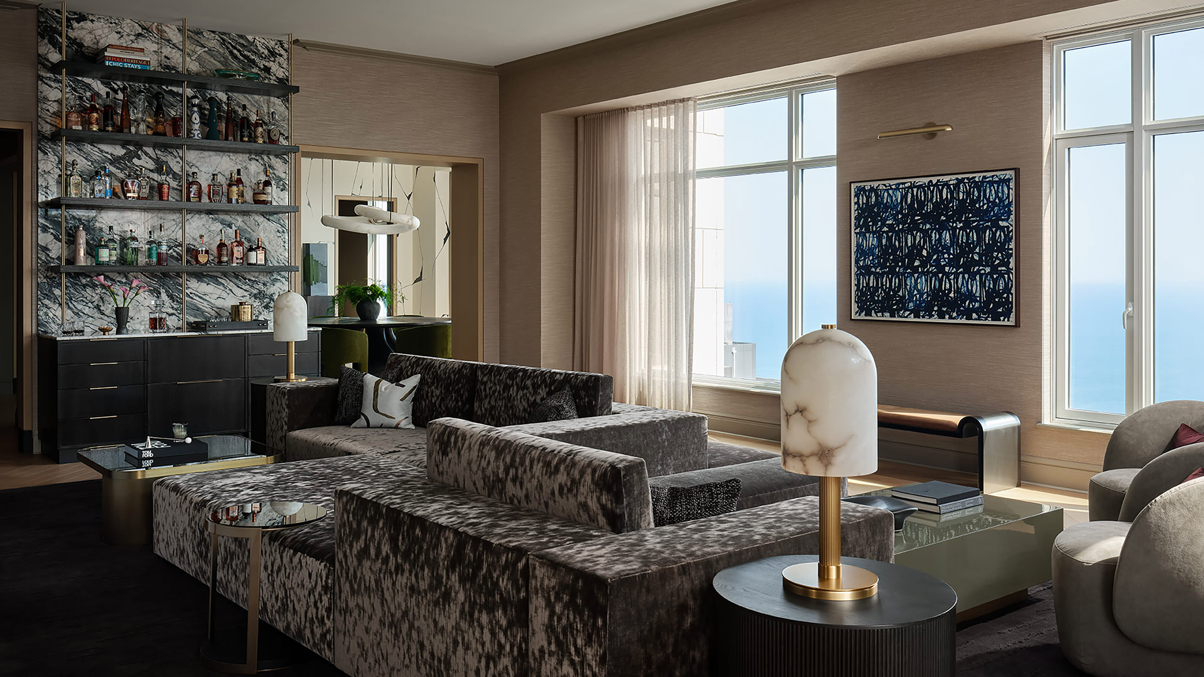 Luxurious living room with plush gray sofas, a marble-topped bar stocked with bottles, large windows with ocean view, and elegant modern decor.
