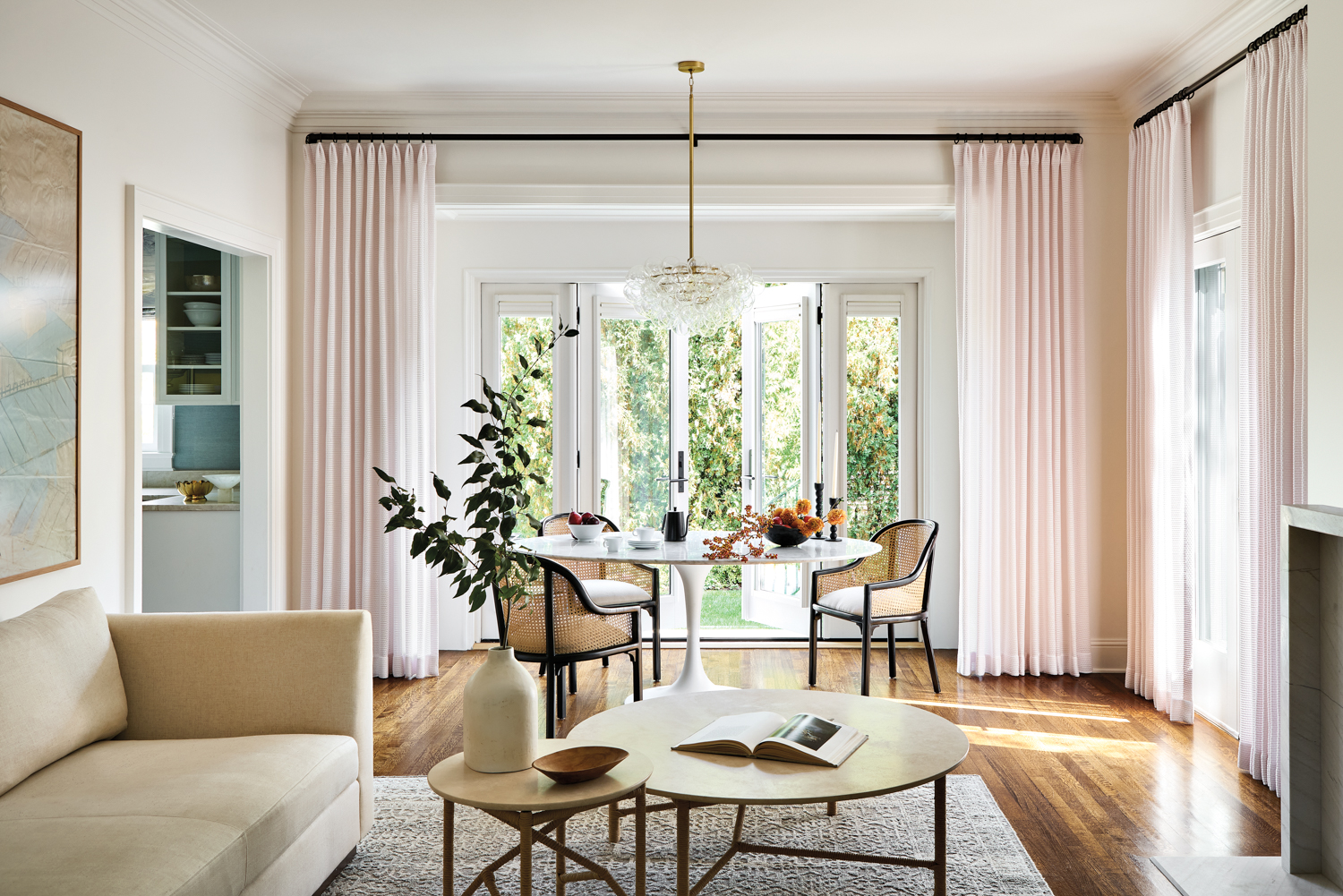 Dining room with pink sheer curtains, a white tulip table and rattan chairs, and a taupe couch by Hayley Kauffmann