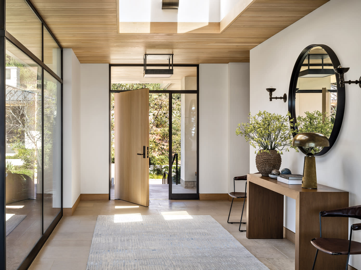 sky light entryway with open pivot door, console table with mirror, lamp and plant, flanked by two chairs