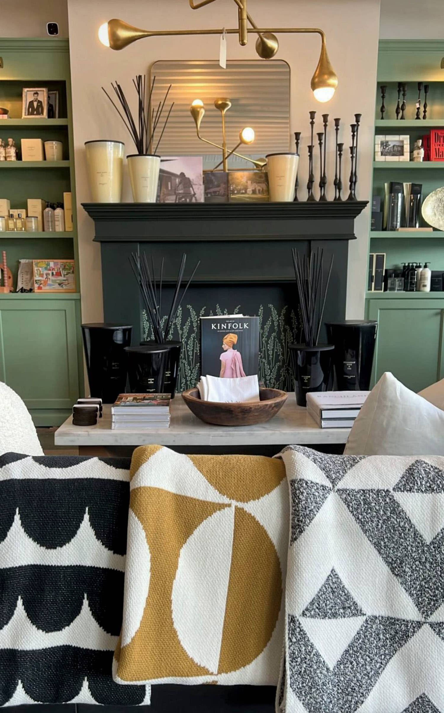 Patterned blankets rest on a sofa across from a fireplace mantle and stocked shelves.
