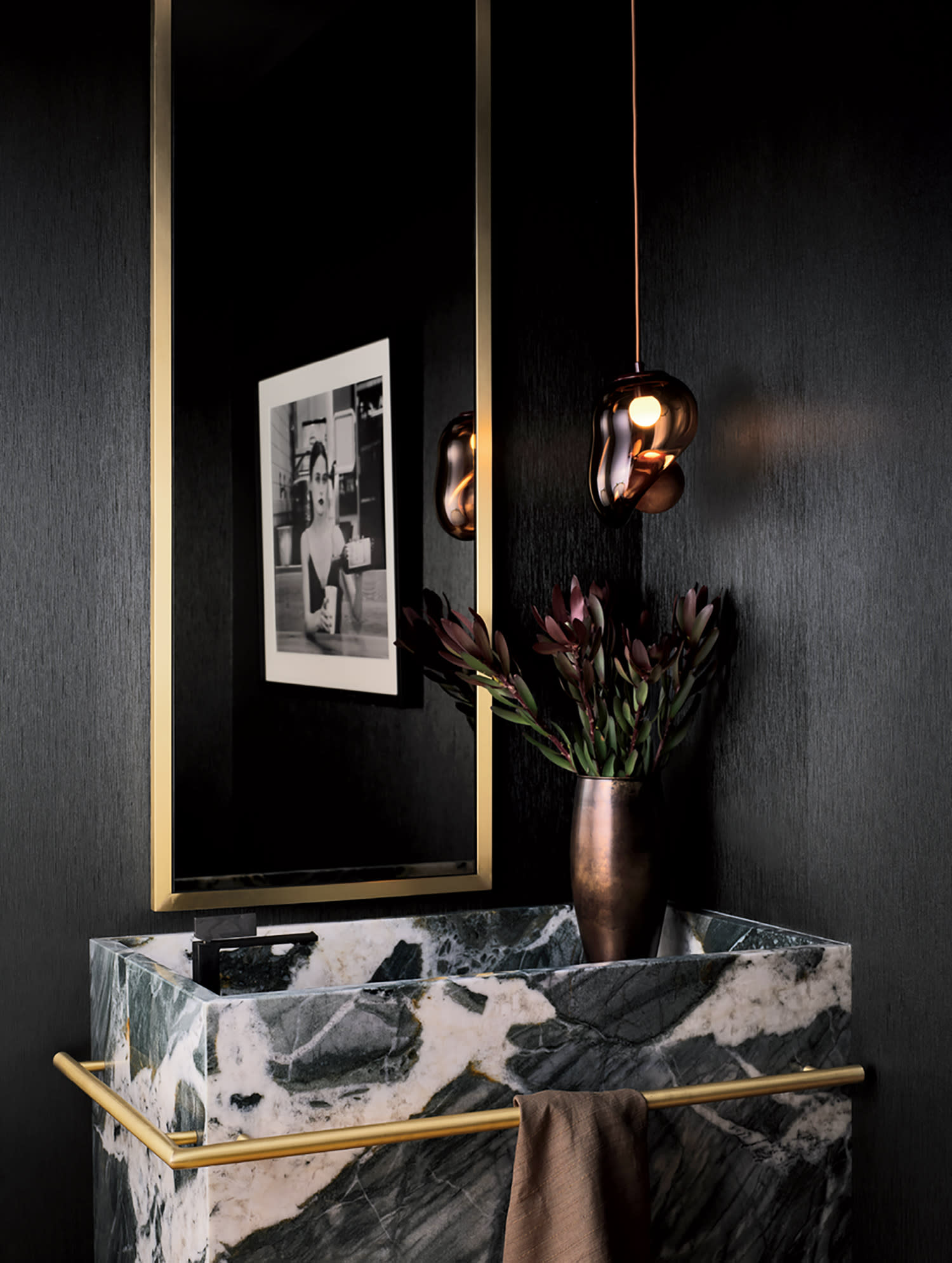 Elegant bathroom with dark walls, a large gold-framed mirror, geometric marble sink, brass towel rail, stylish pendant lamp, and a vase of dark flowers.