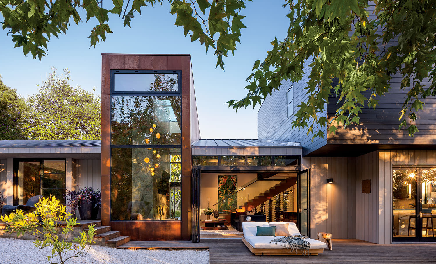 Modern home with a glass facade and large windows, surrounded by greenery. A cozy outdoor seating area on a wooden deck conveys relaxation.
