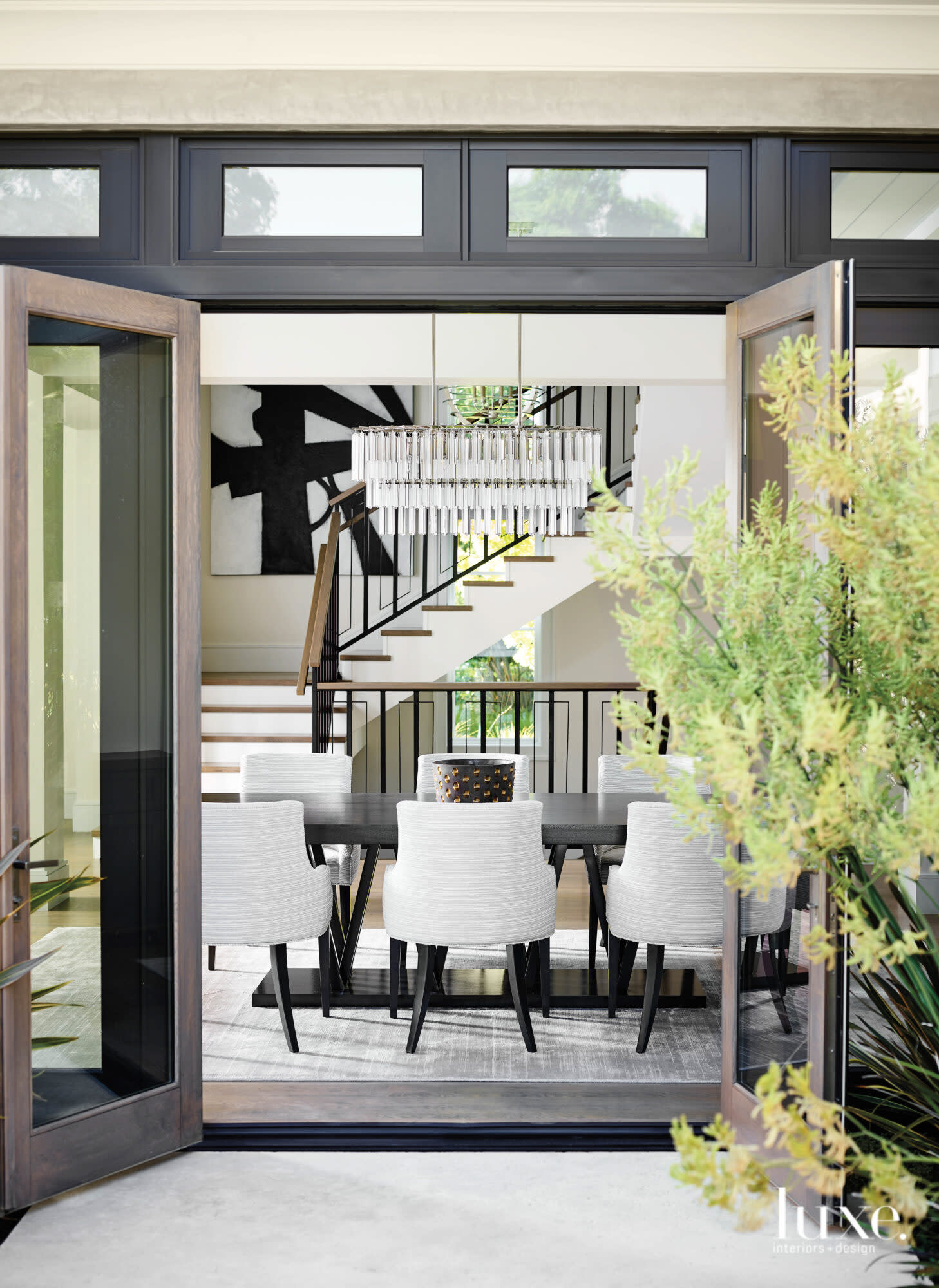Looking from the outside, you see a long dining room table surrounded by white chairs and a long, crystal-studded chandelier.