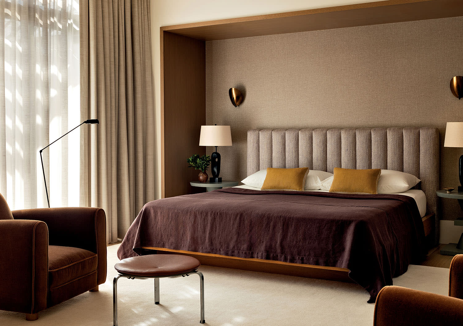 Modern bedroom with a plush bed featuring brown and yellow accents. Flanked by lamps, it includes a cozy chair, footstool, and sheer curtains, evoking warmth.