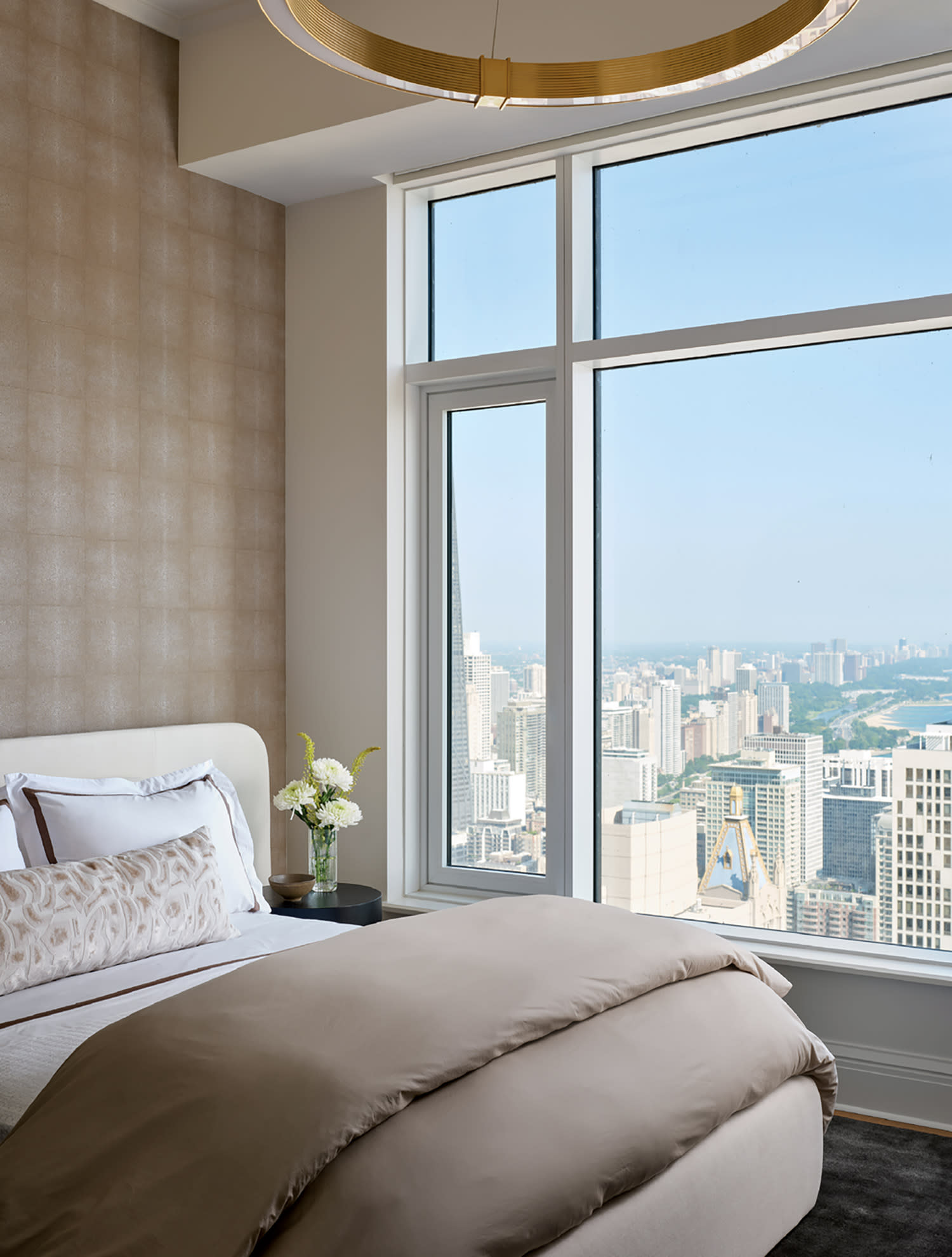 A luxurious bedroom with a large window offering a stunning cityscape view. The bed has beige linens, plush pillows, and a nightstand with flowers.