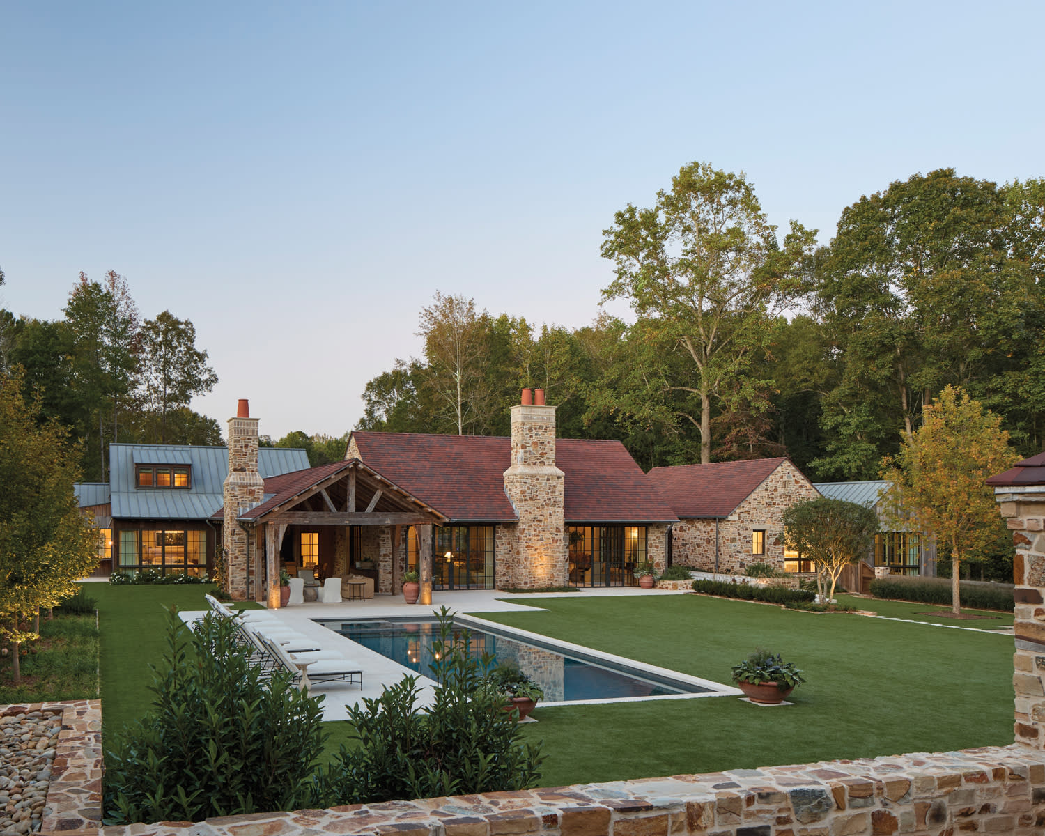 Expansive stone farmhouse with red roof set in lush greenery, featuring two tall chimneys and a rectangular pool, conveys tranquility at sunset.