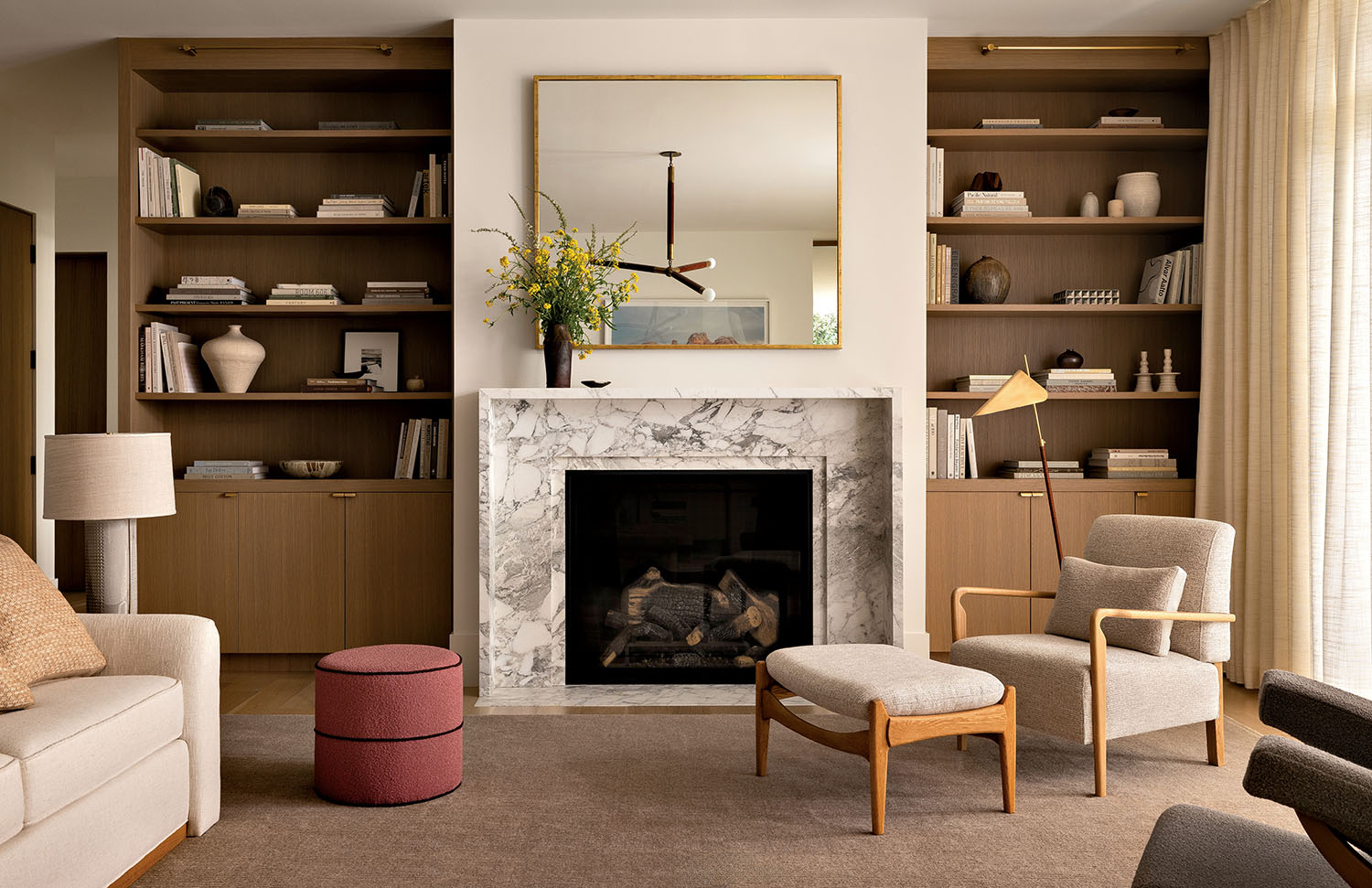Sophisticated living room with a marble fireplace, flanked by wooden bookshelves. Beige furniture, a red ottoman, and a gold mirror create a cozy, modern vibe.
