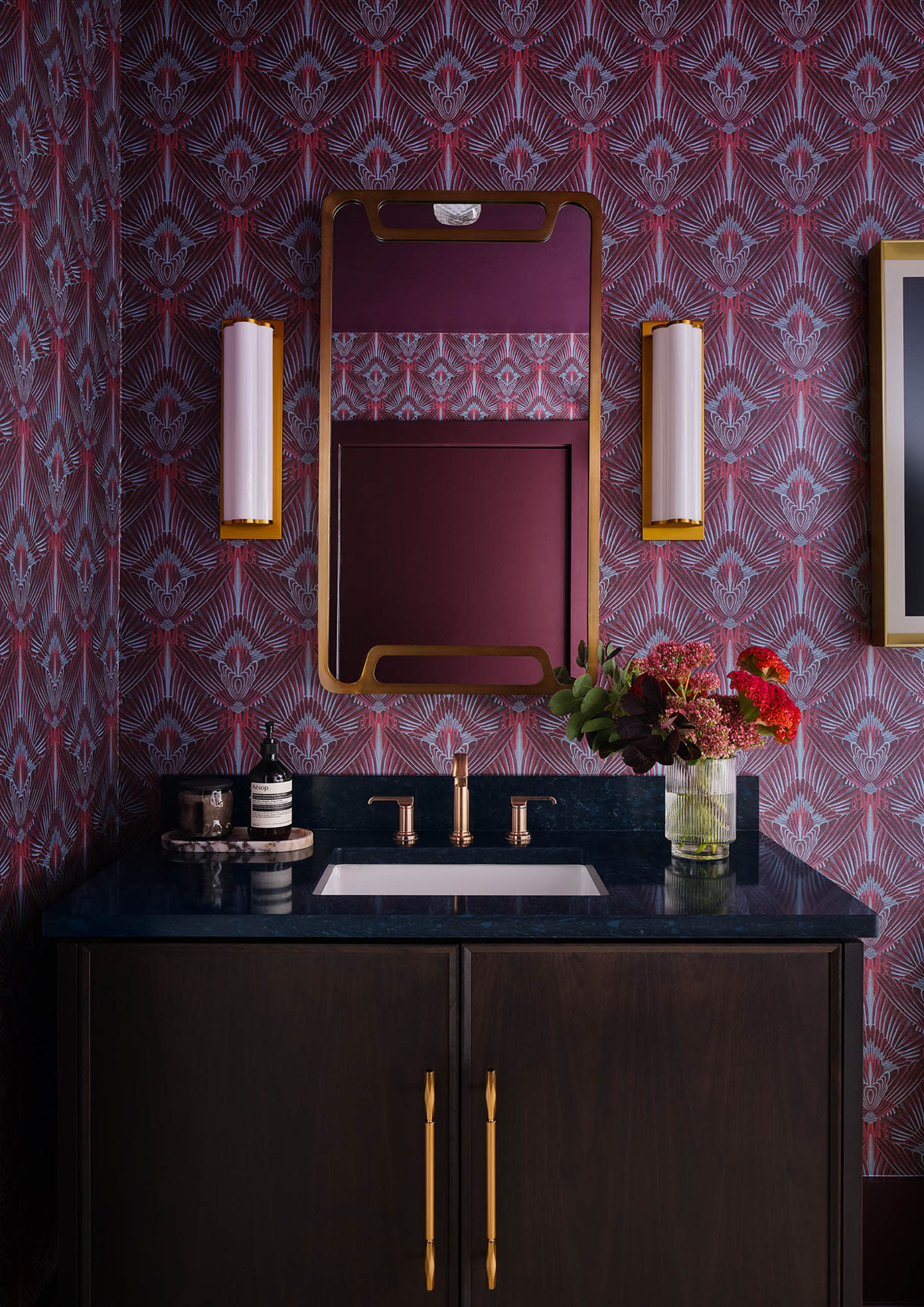 Dark, elegant bathroom with intricate red and blue wallpaper. Features include a gold-framed mirror, two wall lights, a black countertop, and fresh flowers.