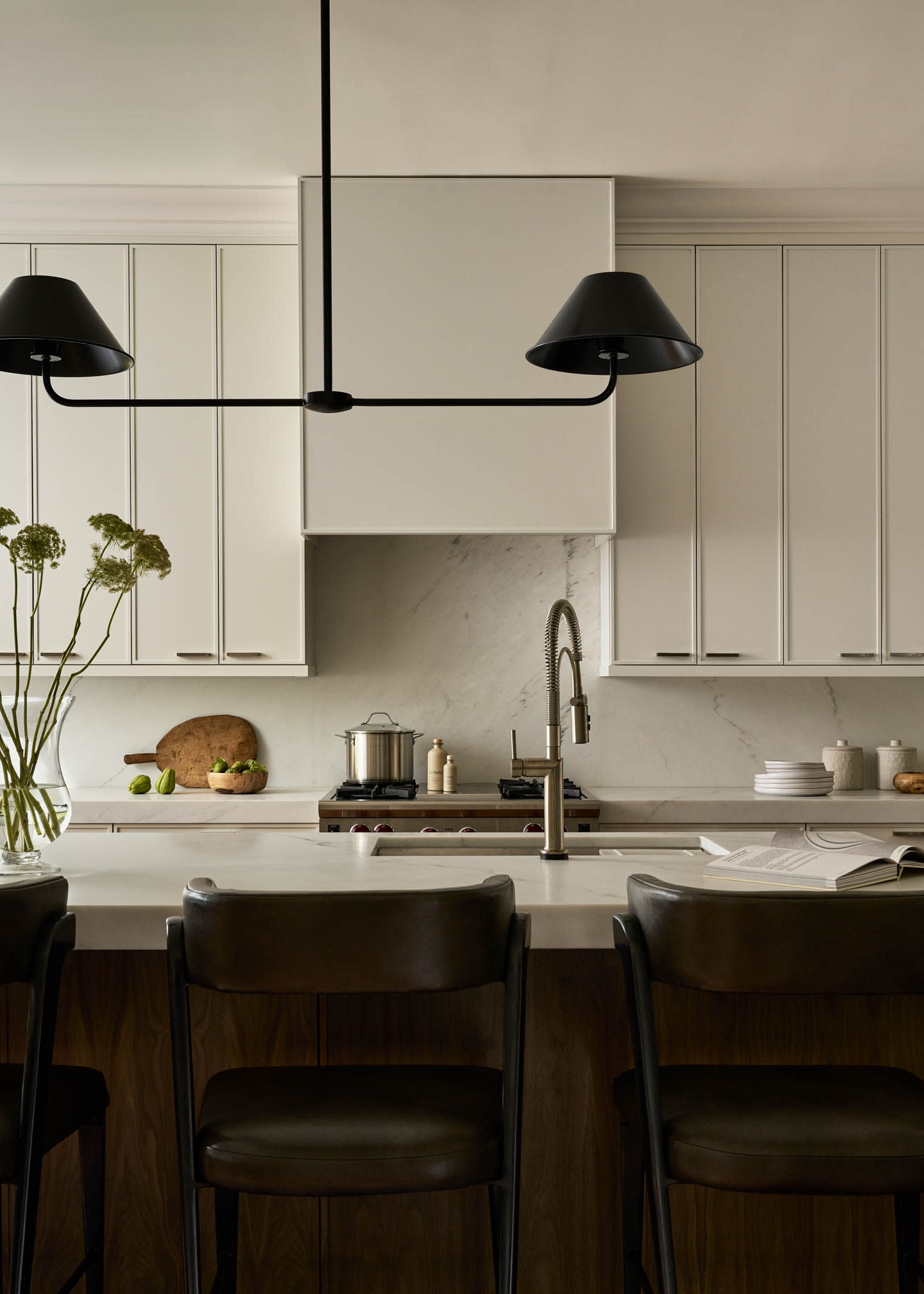 A modern kitchen with sleek white cabinets and marble backsplash. Dark leather chairs line a central island with a minimalist black pendant light above.