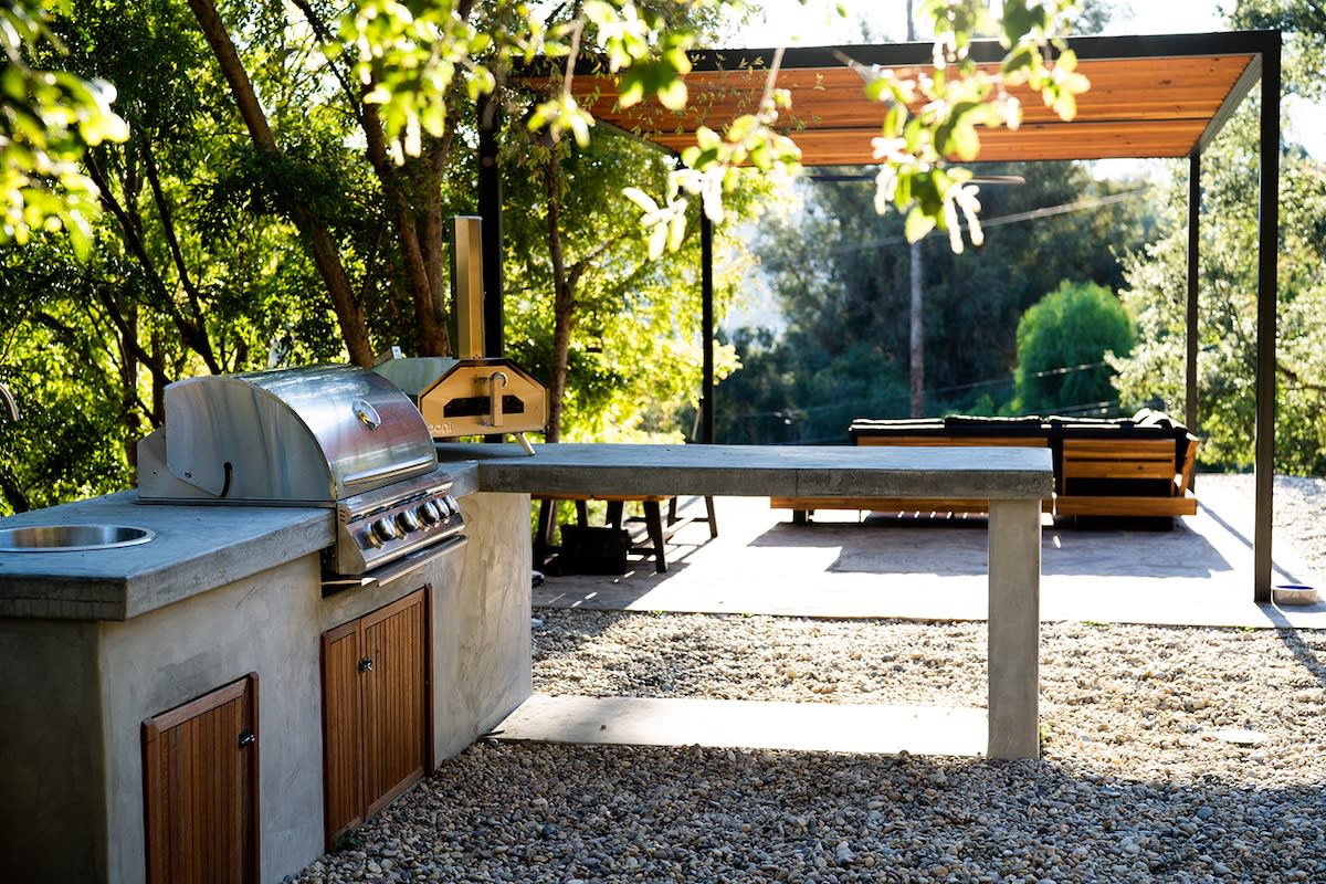 An outdoor kitchen made of concrete, equipped with a grill and a table for dining, surrounded by greenery