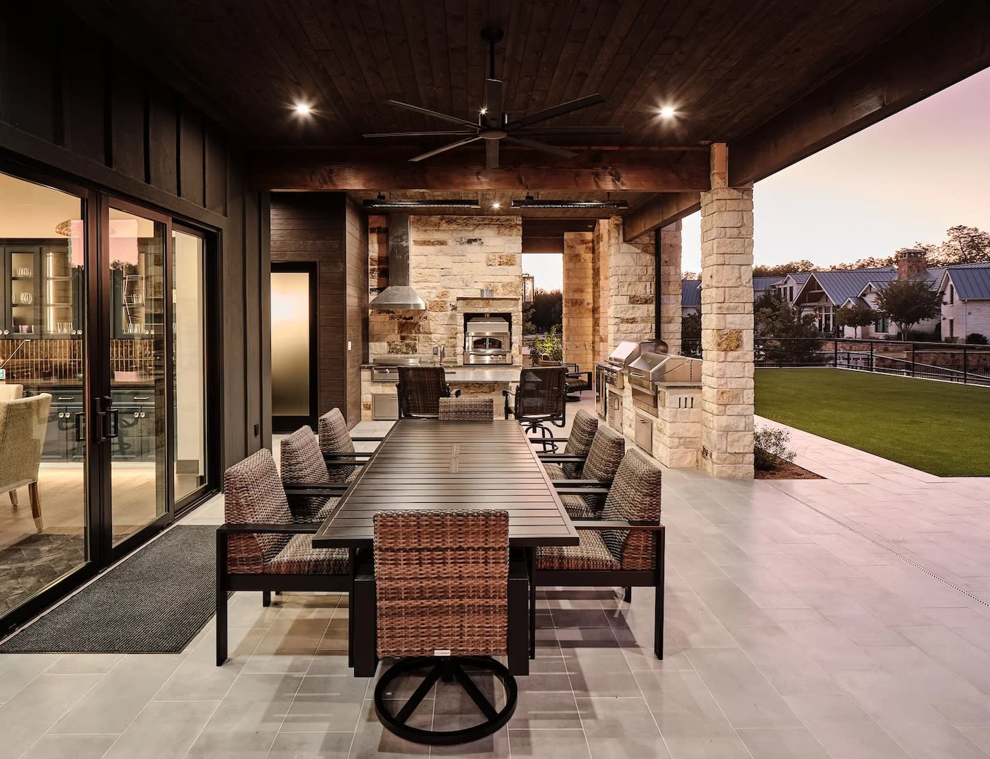 Modern outdoor patio at sunset with a long dining table, wicker chairs, and a stone kitchen with a grill and oven. Cozy, elegant atmosphere.