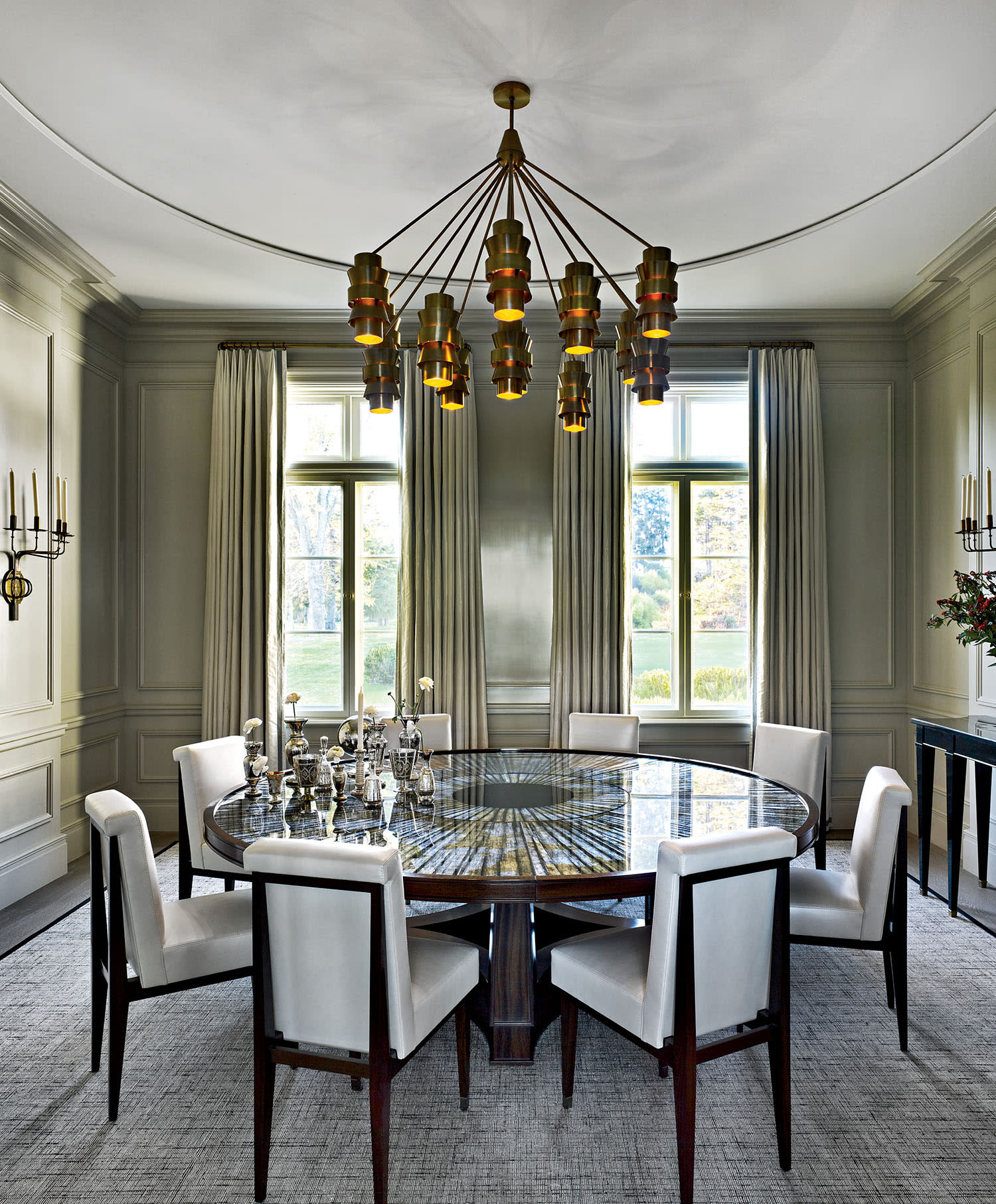 Elegant dining room with a round glass table, white chairs, and a modern chandelier. Large windows with drapes and wall sconces create a sophisticated ambiance.