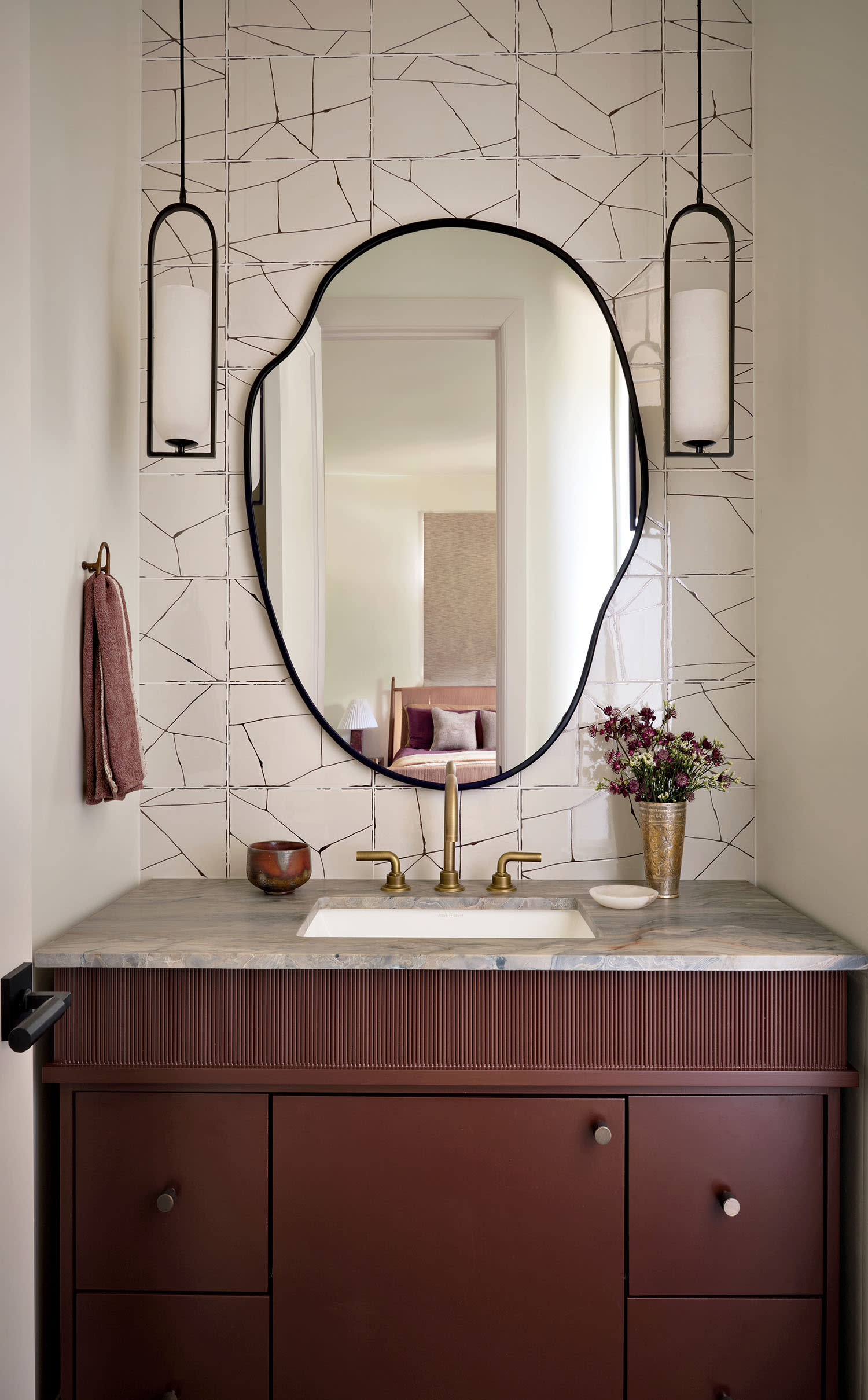 Elegant bathroom with an irregular mirror above a marble-topped, dark wood vanity. Sconces hang beside the mirror; a small vase with flowers adds a cozy touch.