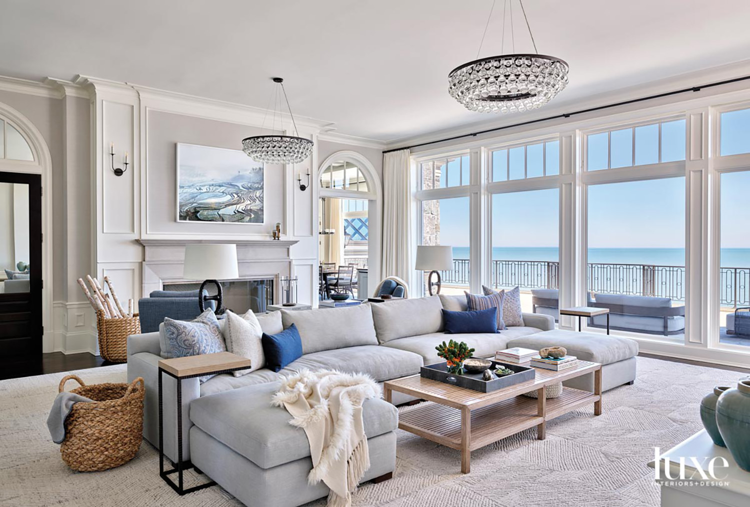 gray living room with large sectional, a fireplace and two chandeliers. The room has floor-to-ceiling windows