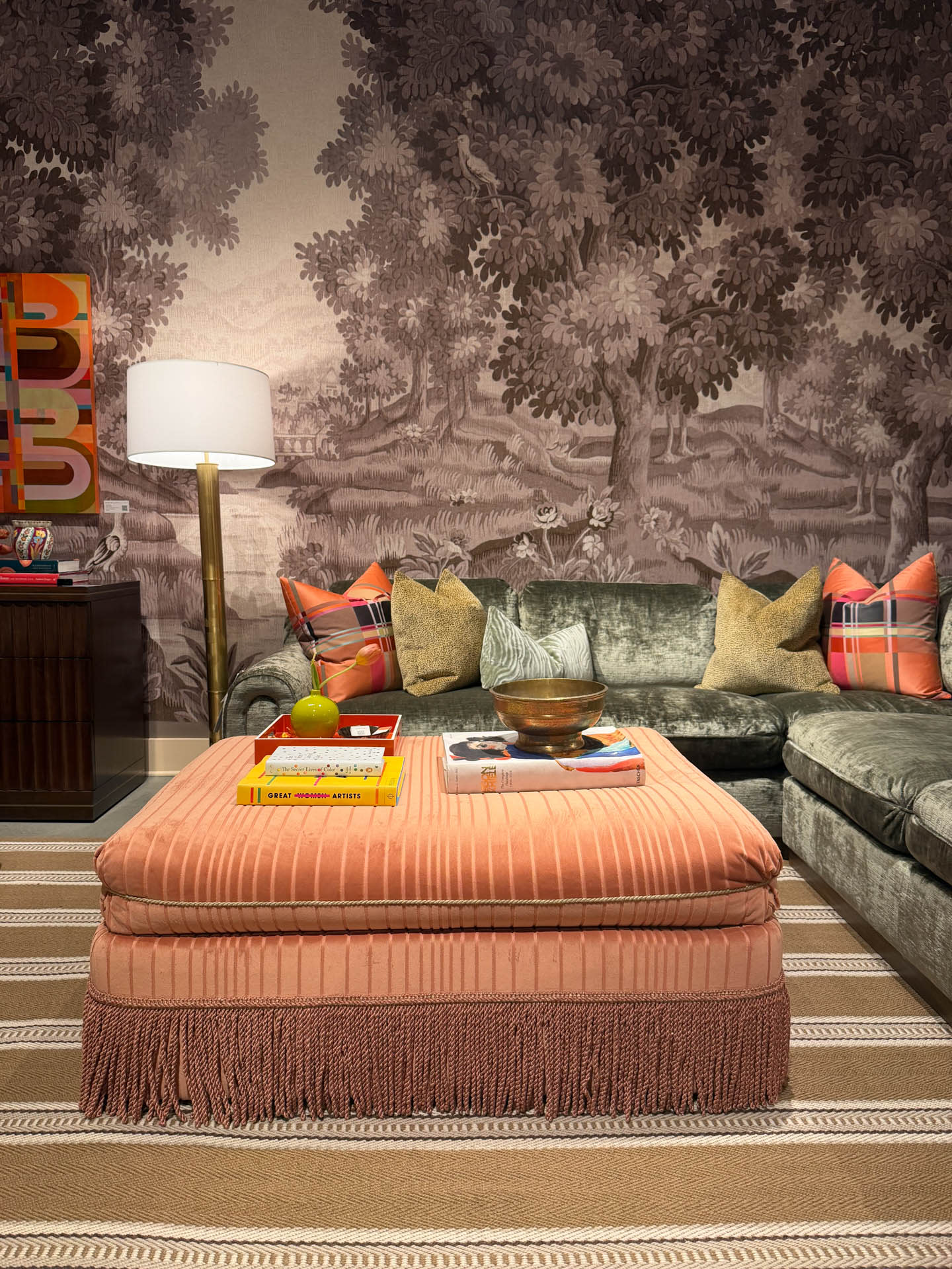 Living room with a pink ottoman, striped rug, and gray sofa adorned with colorful plaid pillows. A forest-themed mural and floor lamp add elegance.