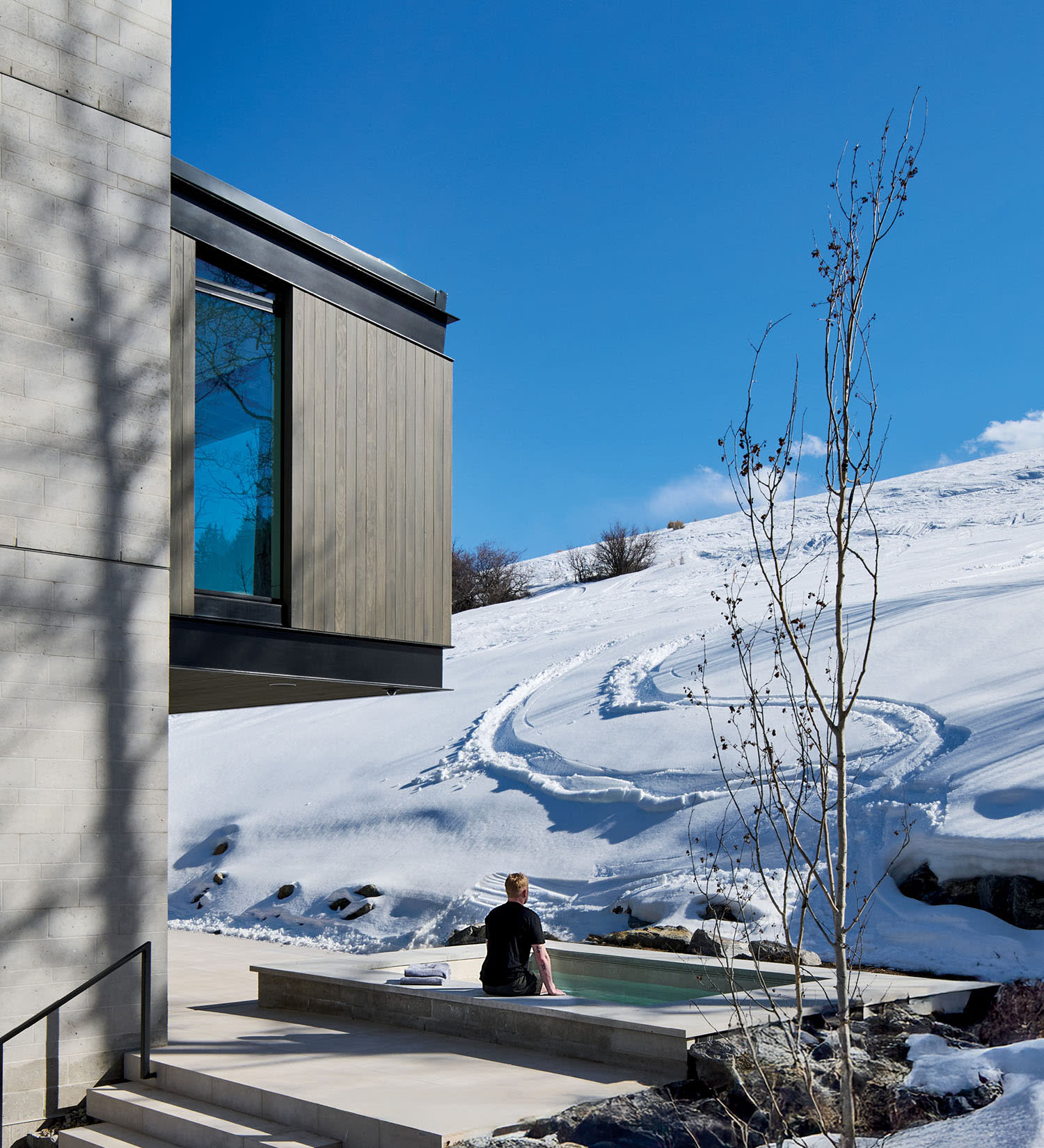 Modern house in snowy landscape under clear blue sky. A person sits by the outdoor hot tub