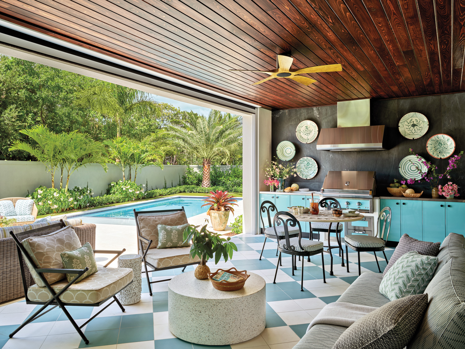 Outdoor kitchen with modern decor features a turquoise kitchen, dining area, and cozy seating. Overlooks a pool surrounded by lush greenery, emitting a relaxed and inviting vibe.