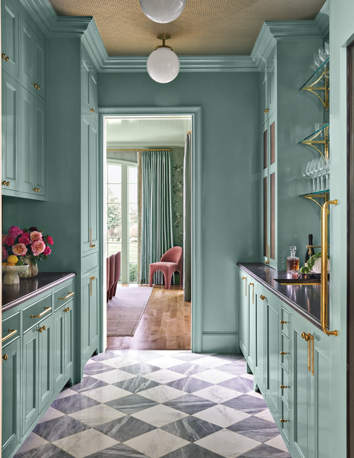 Narrow teal kitchen with checkered floor, brass accents, and globe lights. Doorway reveals a sunlit room with pink chairs and tall windows. Elegant and inviting.