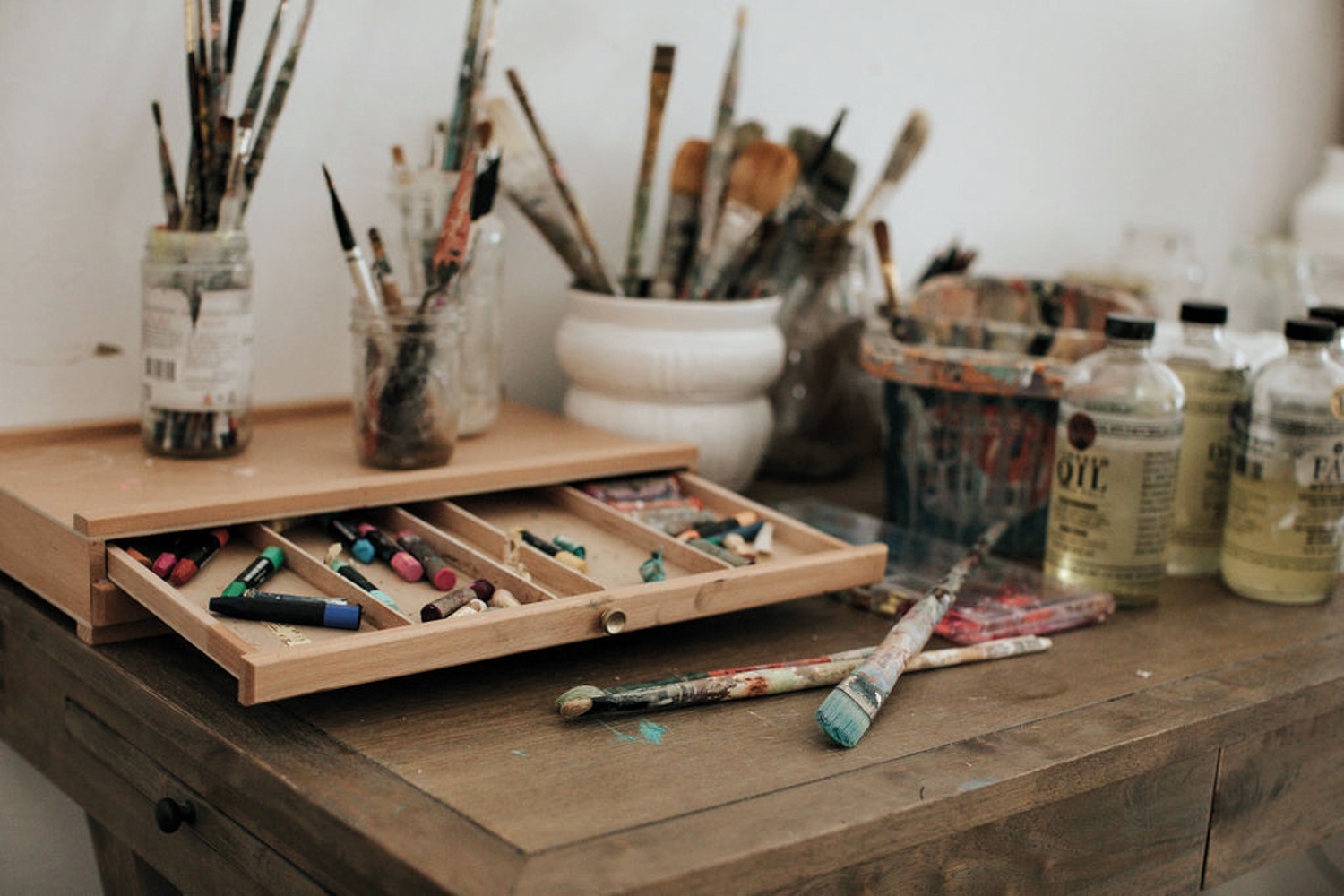 Paint brushes in mason jars and vases beside a draw of pastels on top a wooden table