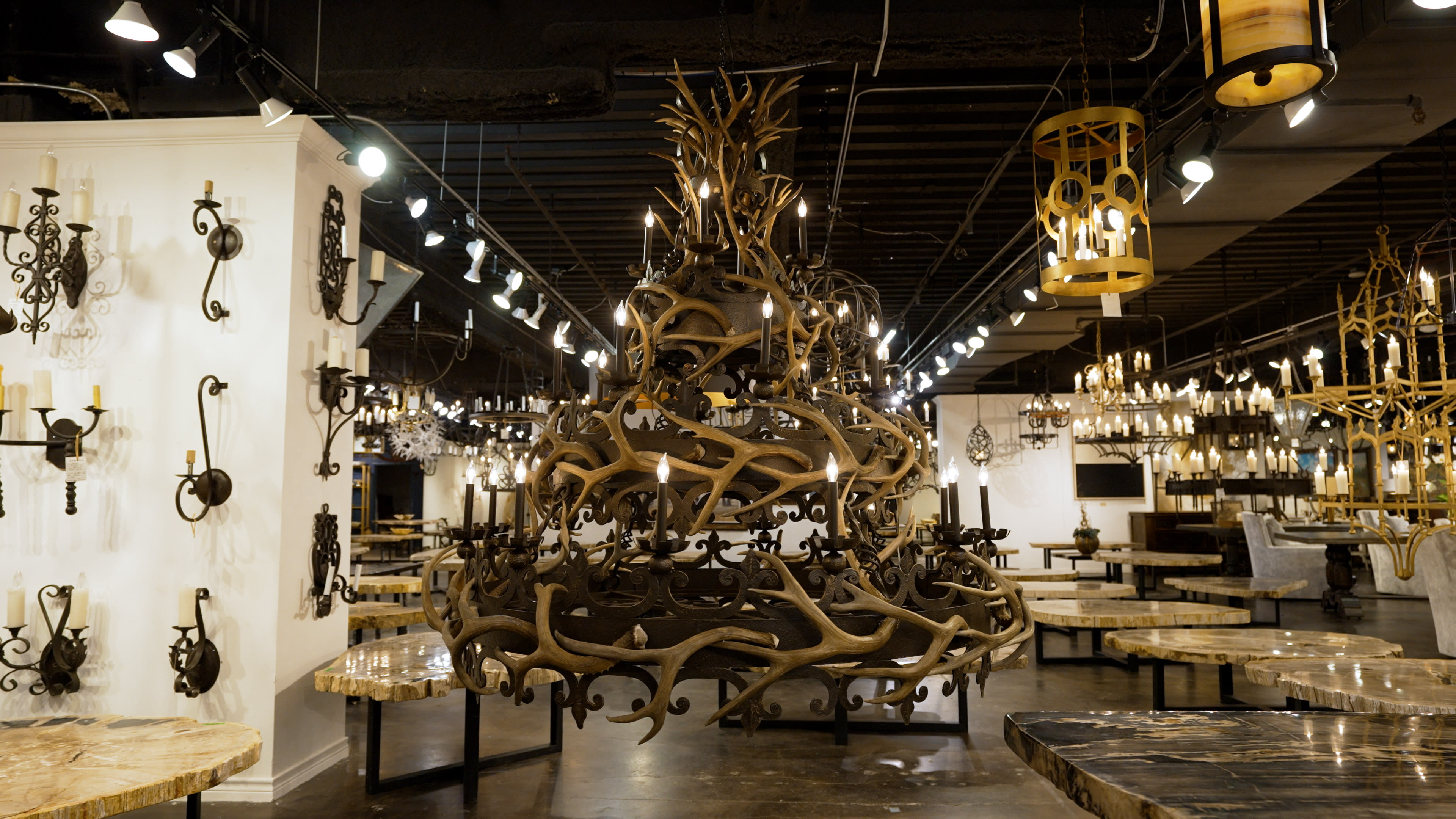 A large, intricate antler chandelier is displayed in a showroom filled with rustic light fixtures.