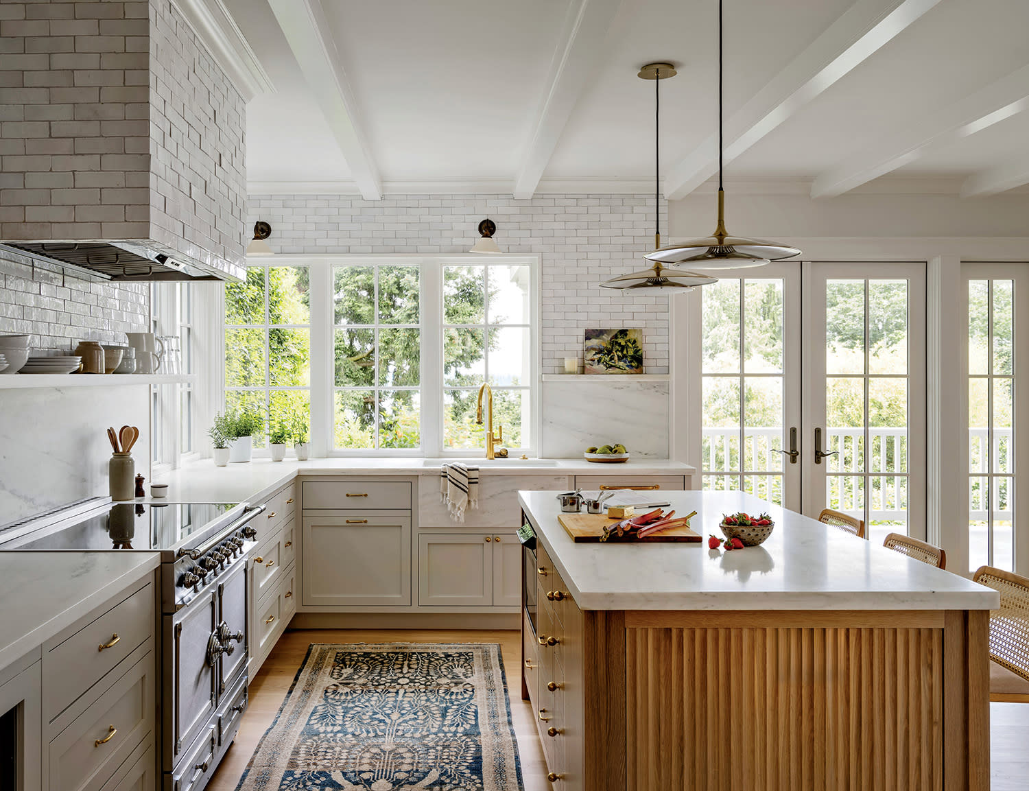 Bright kitchen with white cabinets, a large island, and wood accents. Sunlight streams through large windows, highlighting fresh produce on the counter. Cozy and inviting atmosphere.