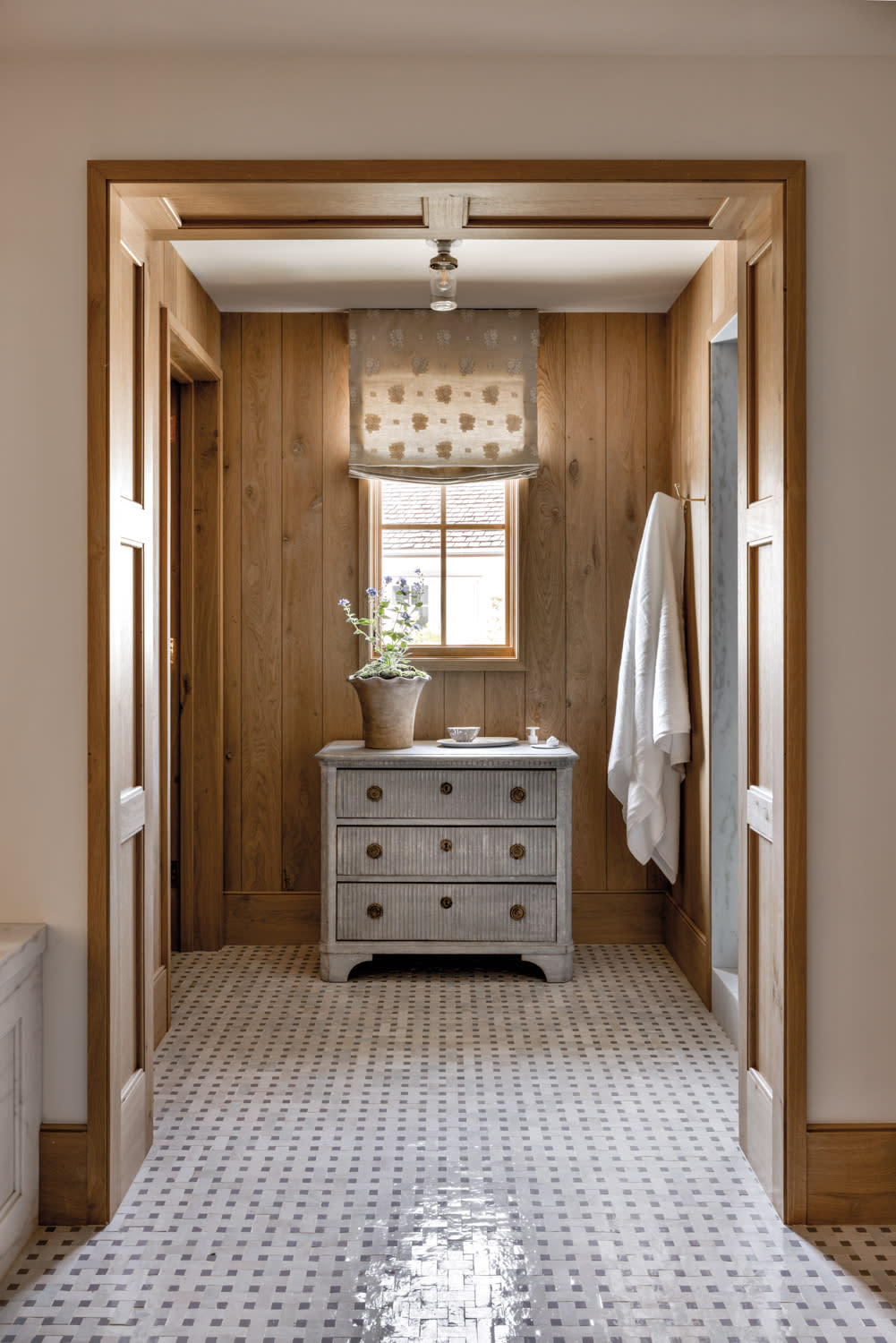 Vintage blue commode in bathroom against oak paneled walls and beneath a window with a floral fabric window treatment by Marie Carson