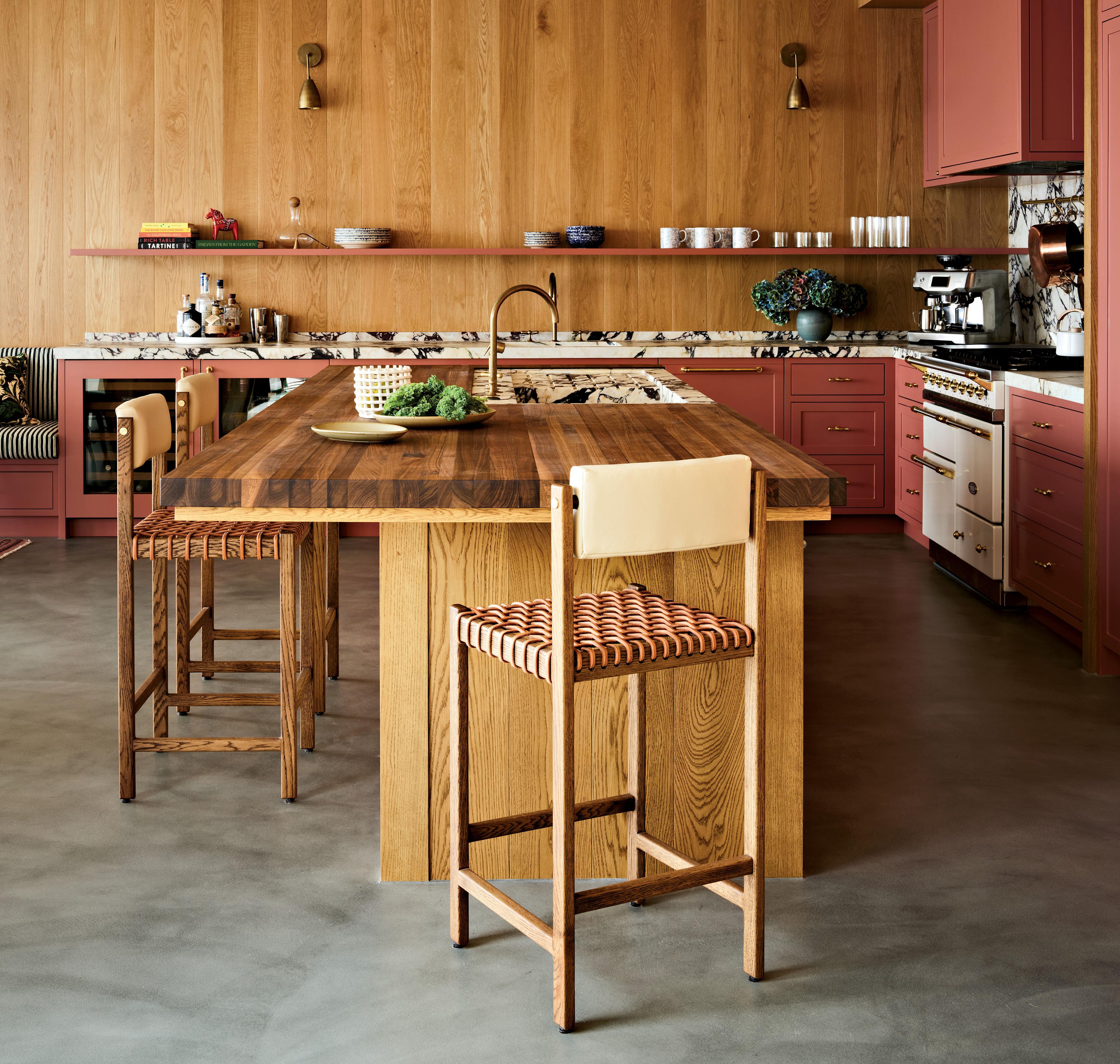 Warm-toned modern kitchen with wooden island, pink cabinets, and sleek appliances. Cozy barstools, open shelving, and decorative plants create an inviting atmosphere.
