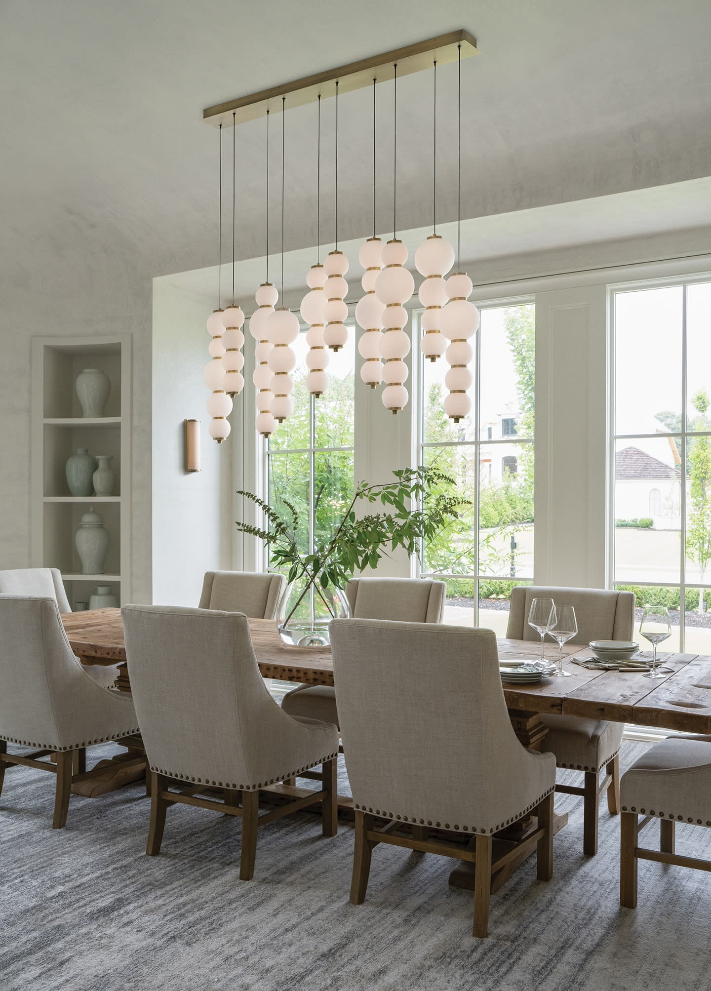 Elegant dining room with a wooden table, beige upholstered chairs, modern cluster pendant lights, large windows, and green plant centerpiece.