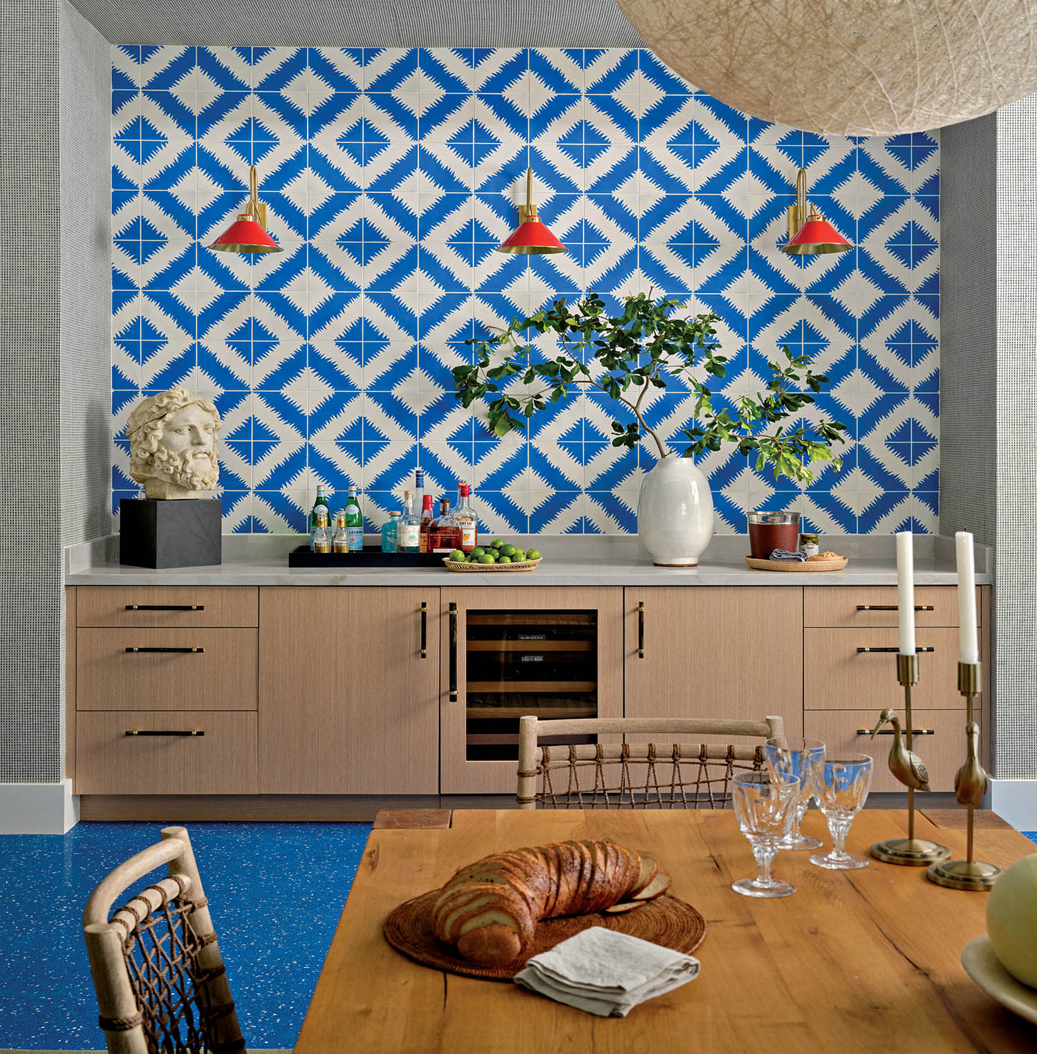 Dining area with a vibrant blue and white geometric wall, red pendant lights, a wooden table set with croissants, and modern decor creating a lively, artistic ambiance.