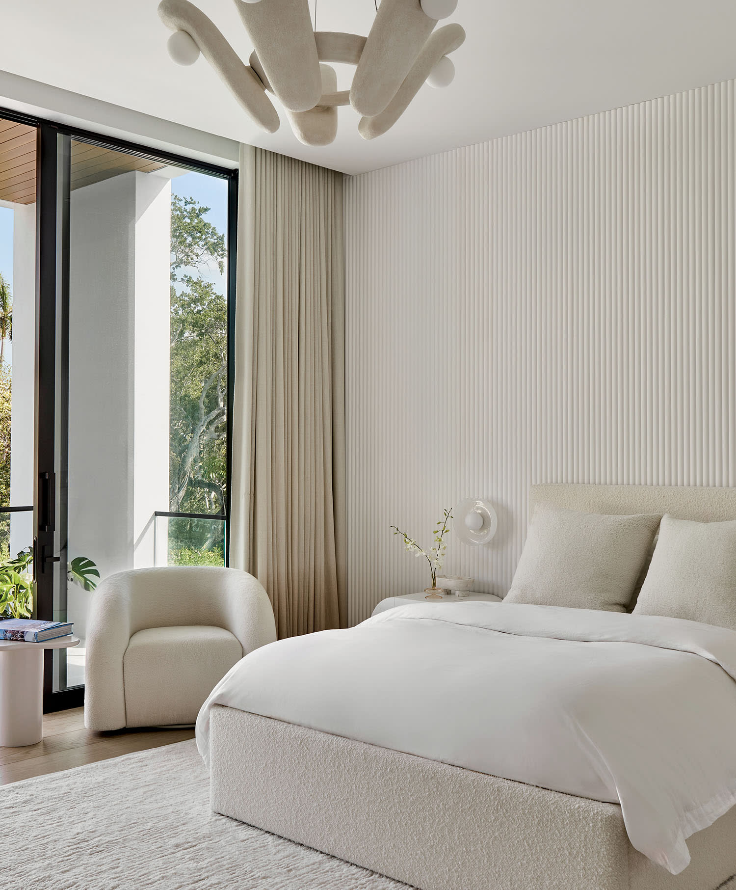 Minimalist bedroom with a plush bed, light beige armchair, and unique abstract chandelier. Floor-to-ceiling window and neutral tones create a serene atmosphere.